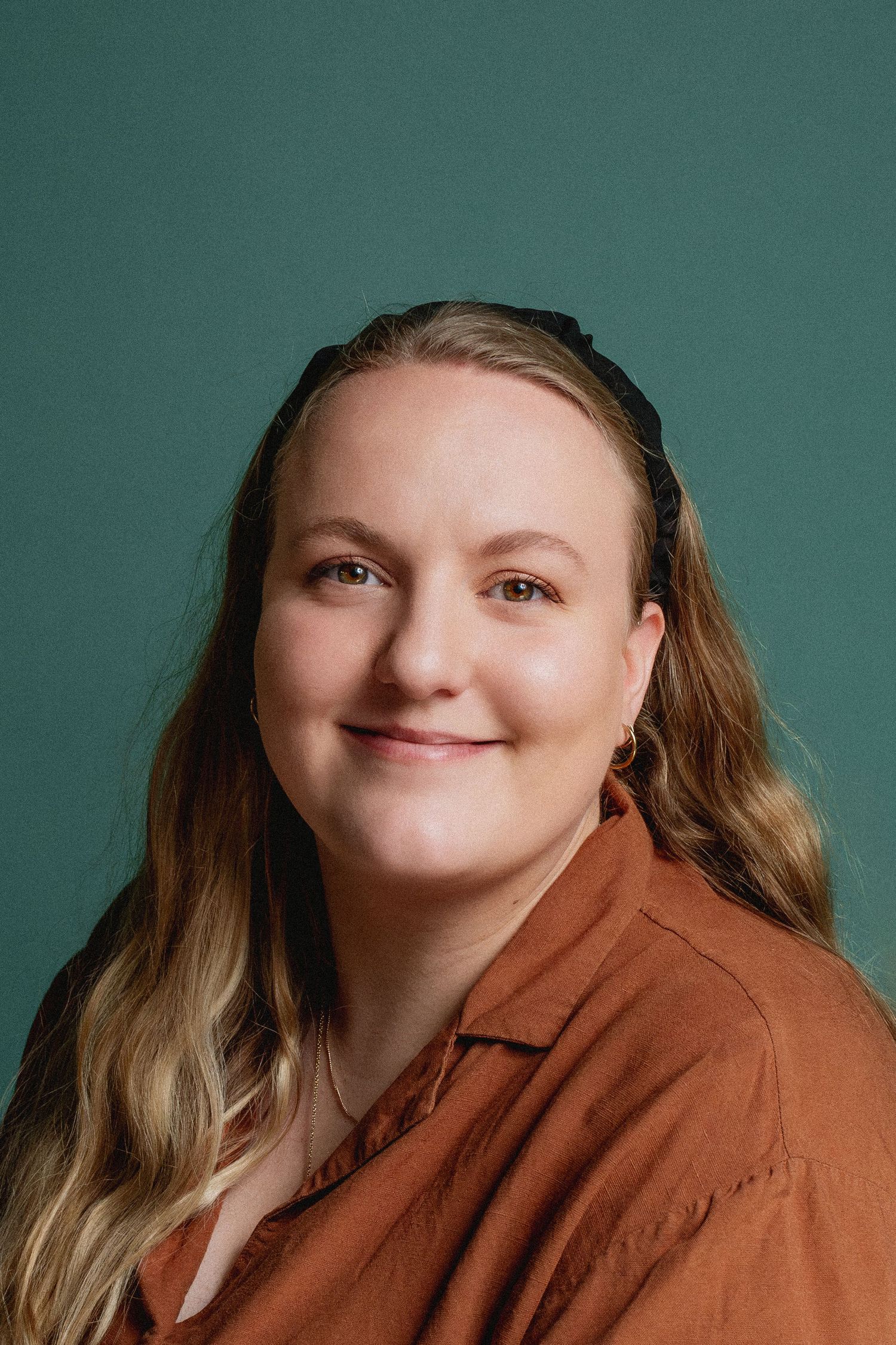 Professional headshot of a person in a rust-colored blazer against a teal background smiling warmly at the camera.