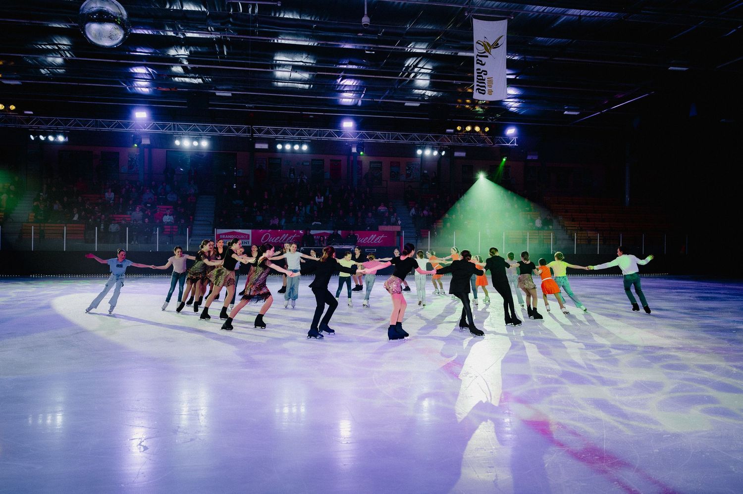 Ice skaters perform in a synchronized routine on a dark illuminated rink with colorful spotlights shining down.
