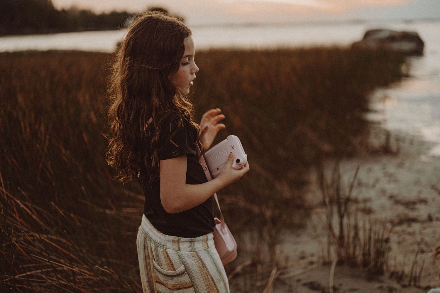 Un enfant se tient parmi les hautes herbes au coucher du soleil, tenant un appareil photo près de l’eau.
