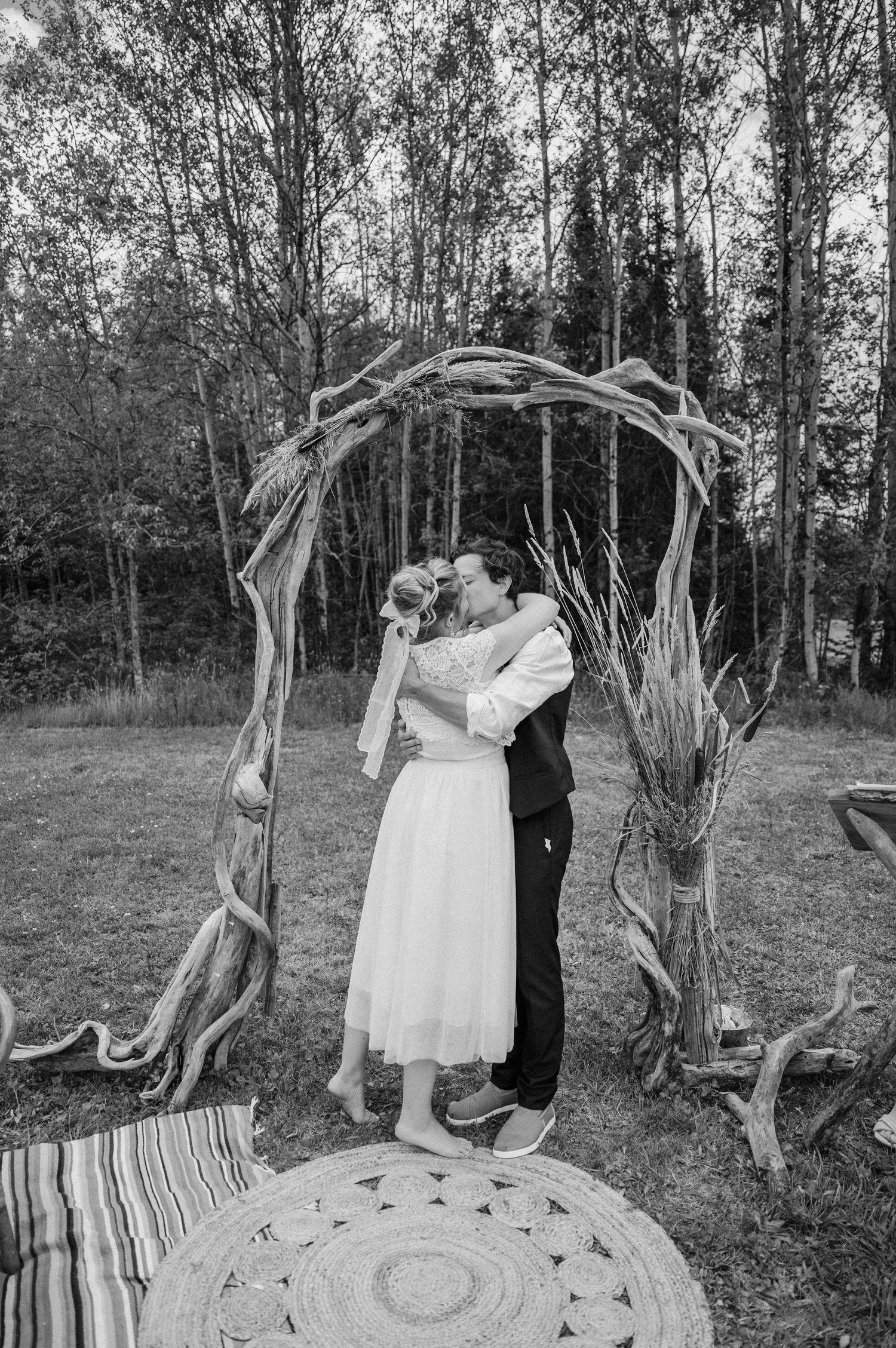 A couple embraces under a driftwood arch in a black and white forest setting.