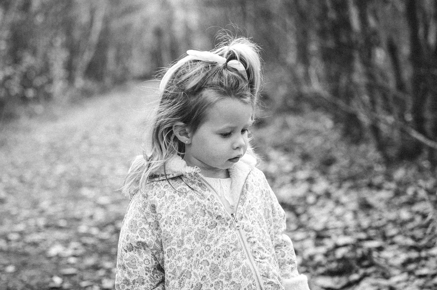 Black and white portrait of a small child in a floral dress walking through autumn woods.