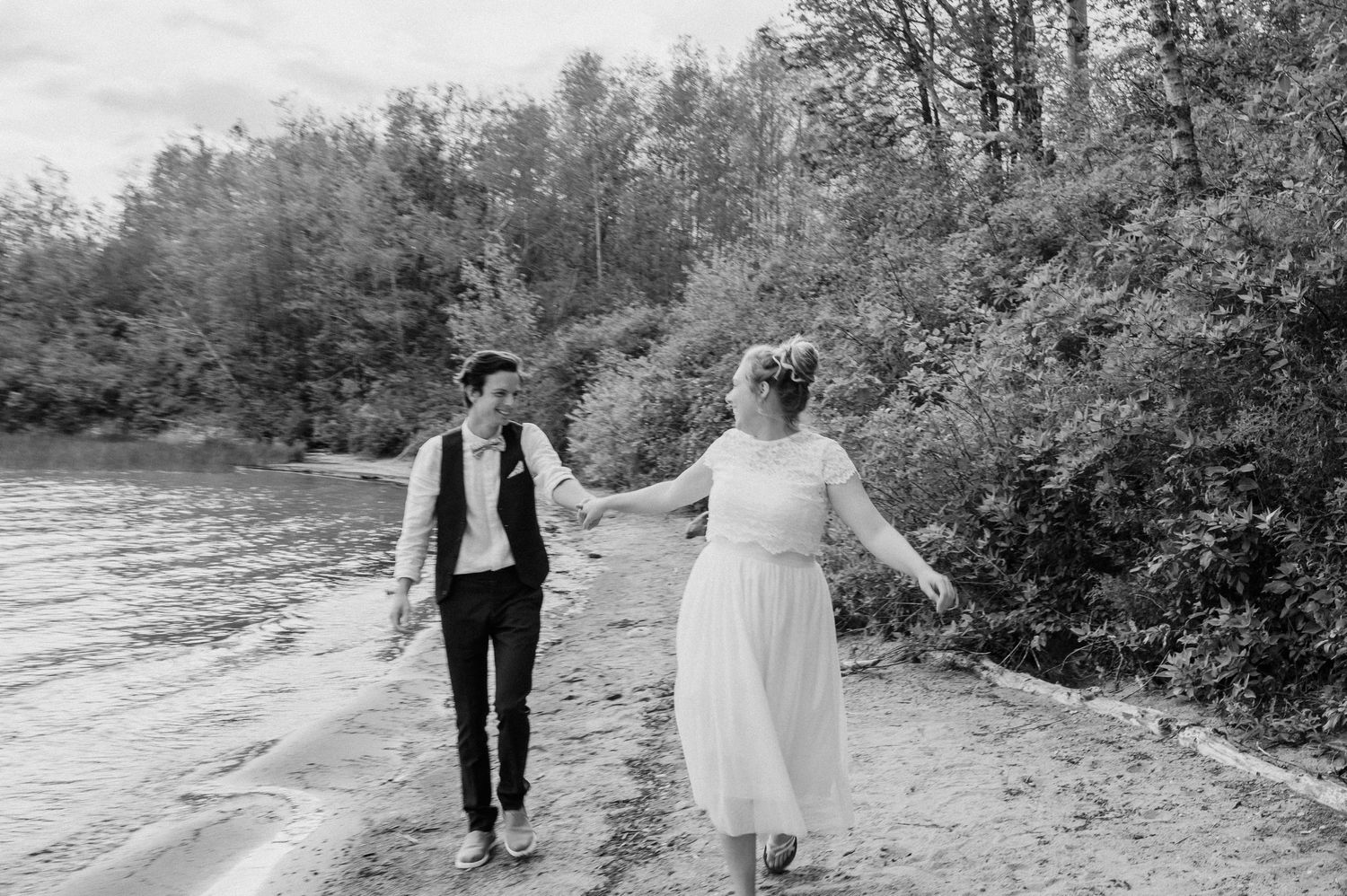 A couple walks hand in hand along a riverbank in black and white.
