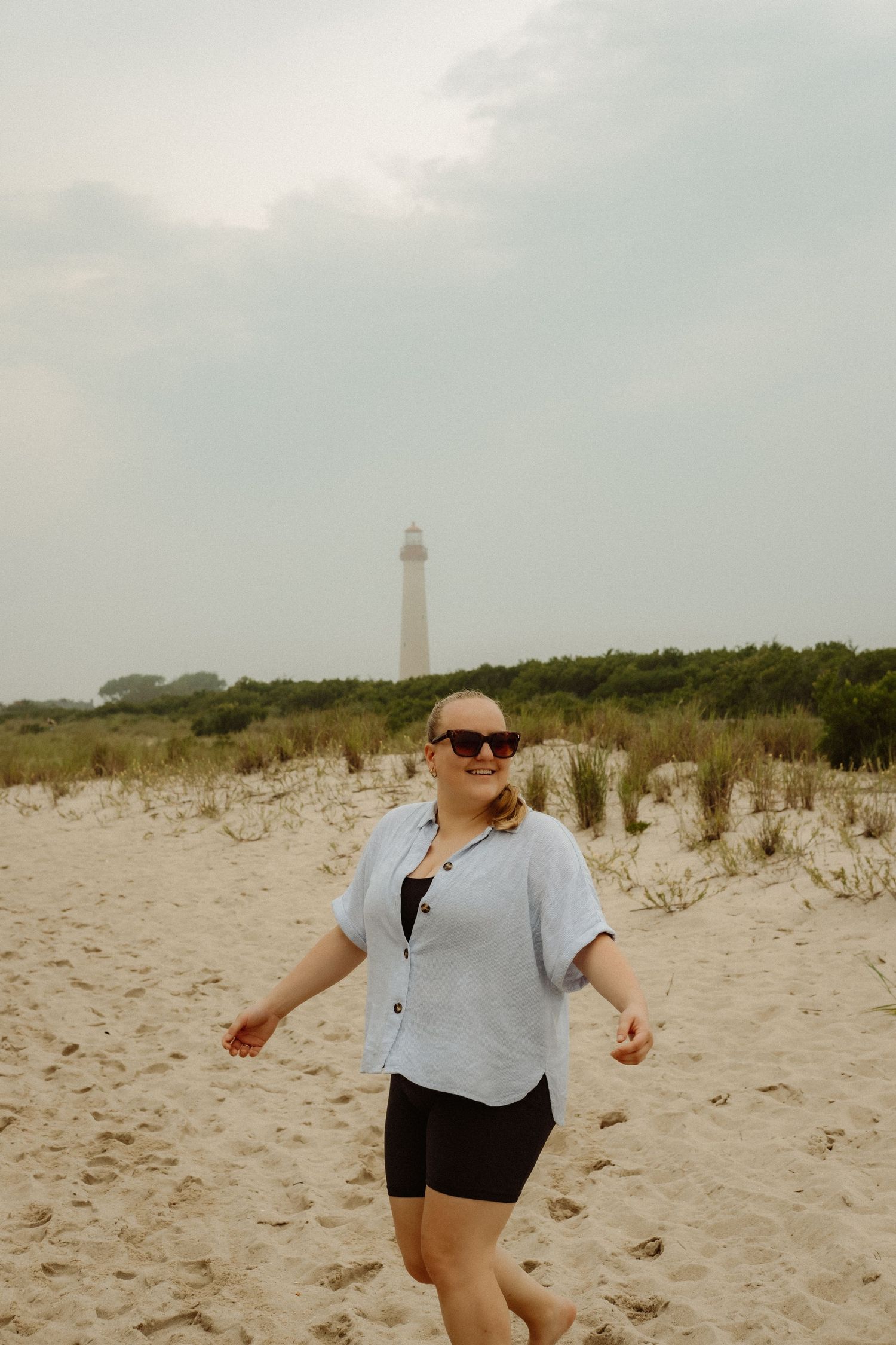 Une silhouette en gilet léger et short se tient sur une plage de sable, avec un phare visible dans le brouillard en arrière-plan.