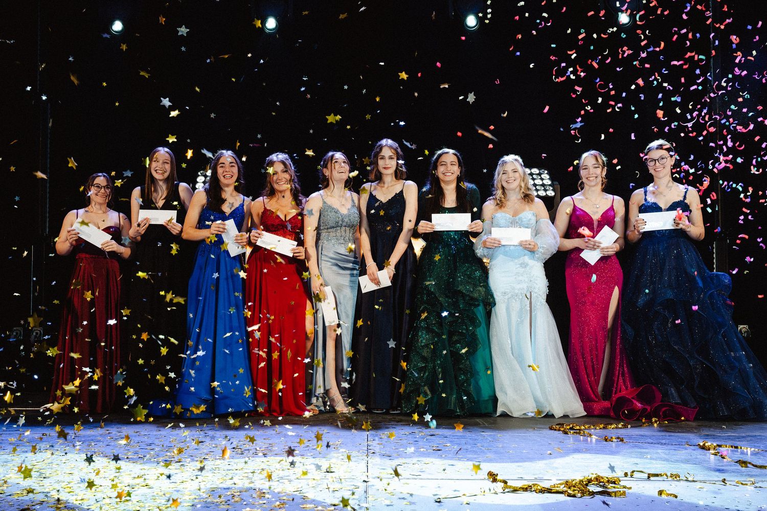 A group in formal gowns and evening wear poses together as confetti falls during a festive celebration.