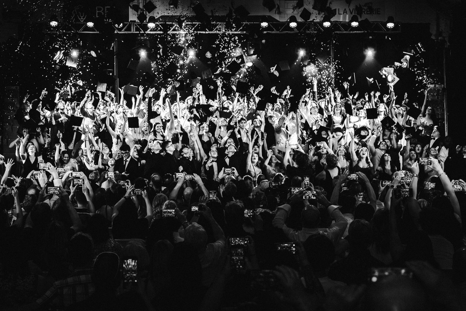 A black and white concert photo shows an energetic crowd with raised hands at a packed music venue during a performance.
