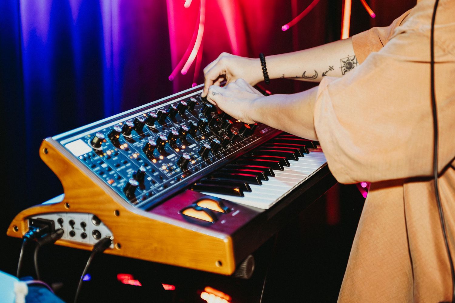 Hand adjusting knobs on a vintage synthesizer keyboard with colorful red and blue stage lighting in the background.