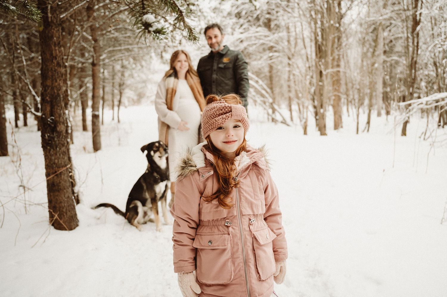 A winter family portrait in a snowy forest with a dog, featuring someone in a pink coat in the foreground.