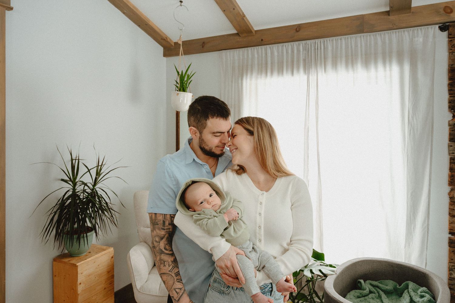 Family relaxes together in bright living room with houseplants near window with sheer curtains.