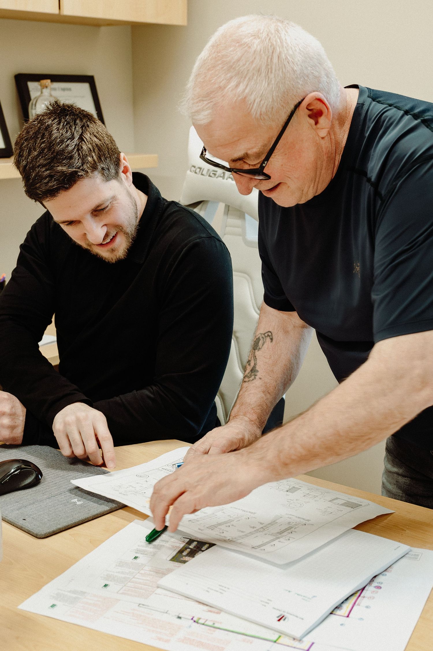 Two people reviewing and discussing architectural plans and documents while working at a wooden desk together.