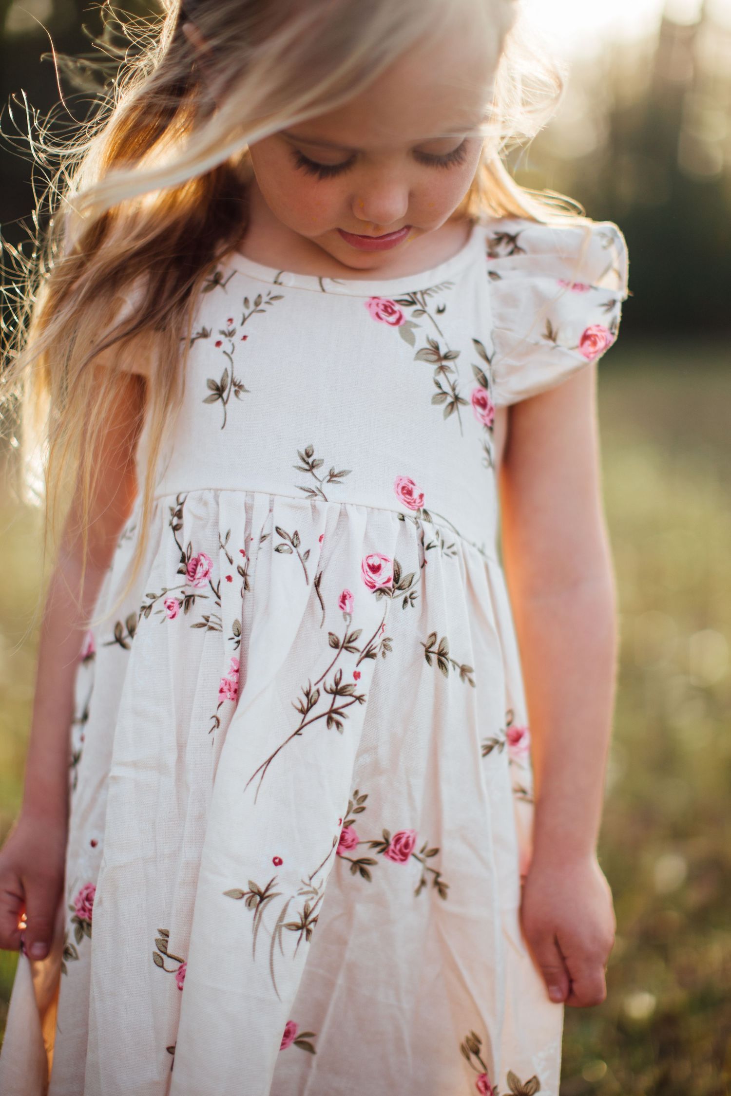 Young girl in a white floral dress with pink roses stands in golden sunlit field with flowing blonde hair.