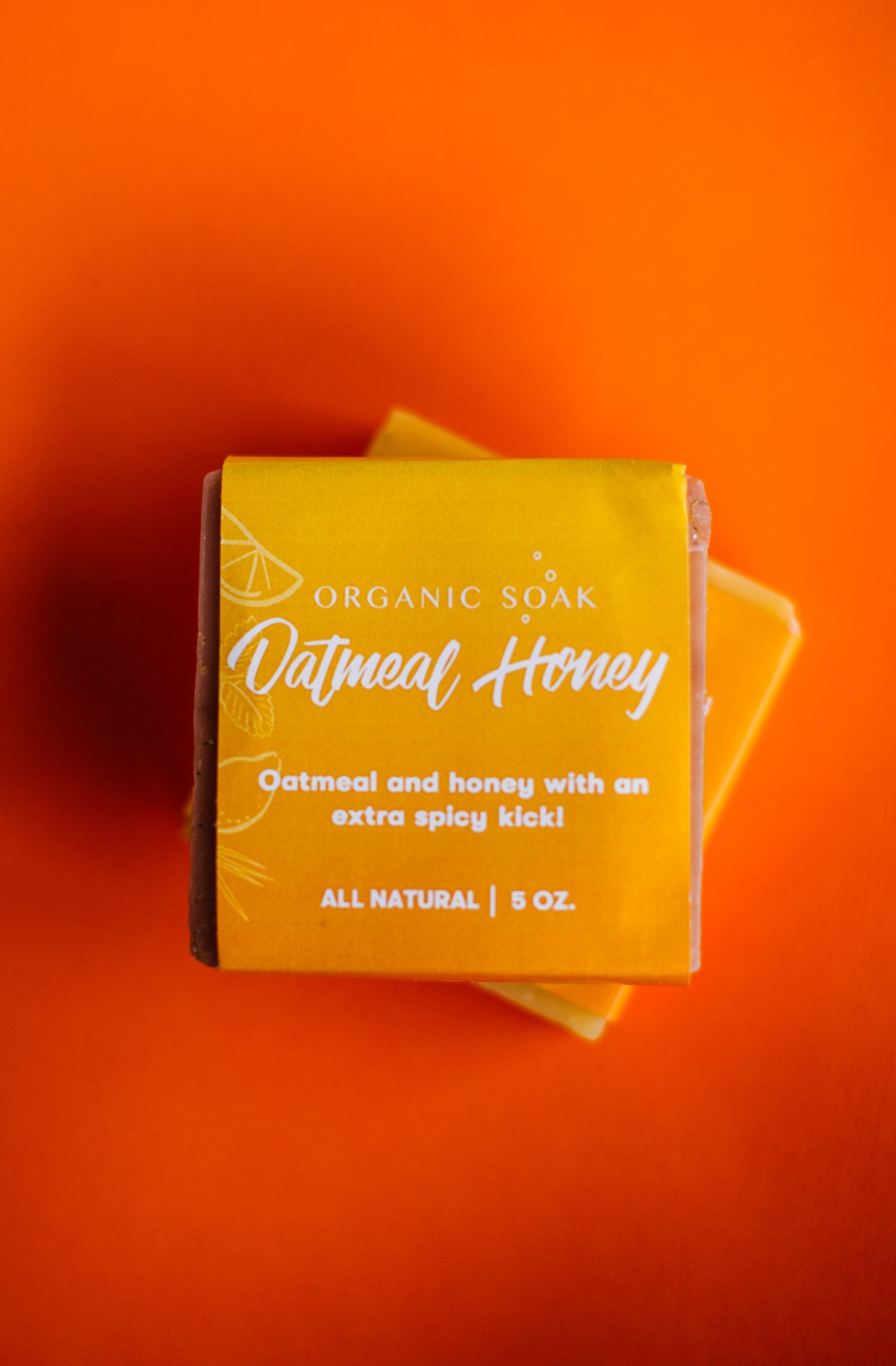 A yellow organic soap bar with 'Oatmeal and Honey' text displayed against a vibrant orange background.