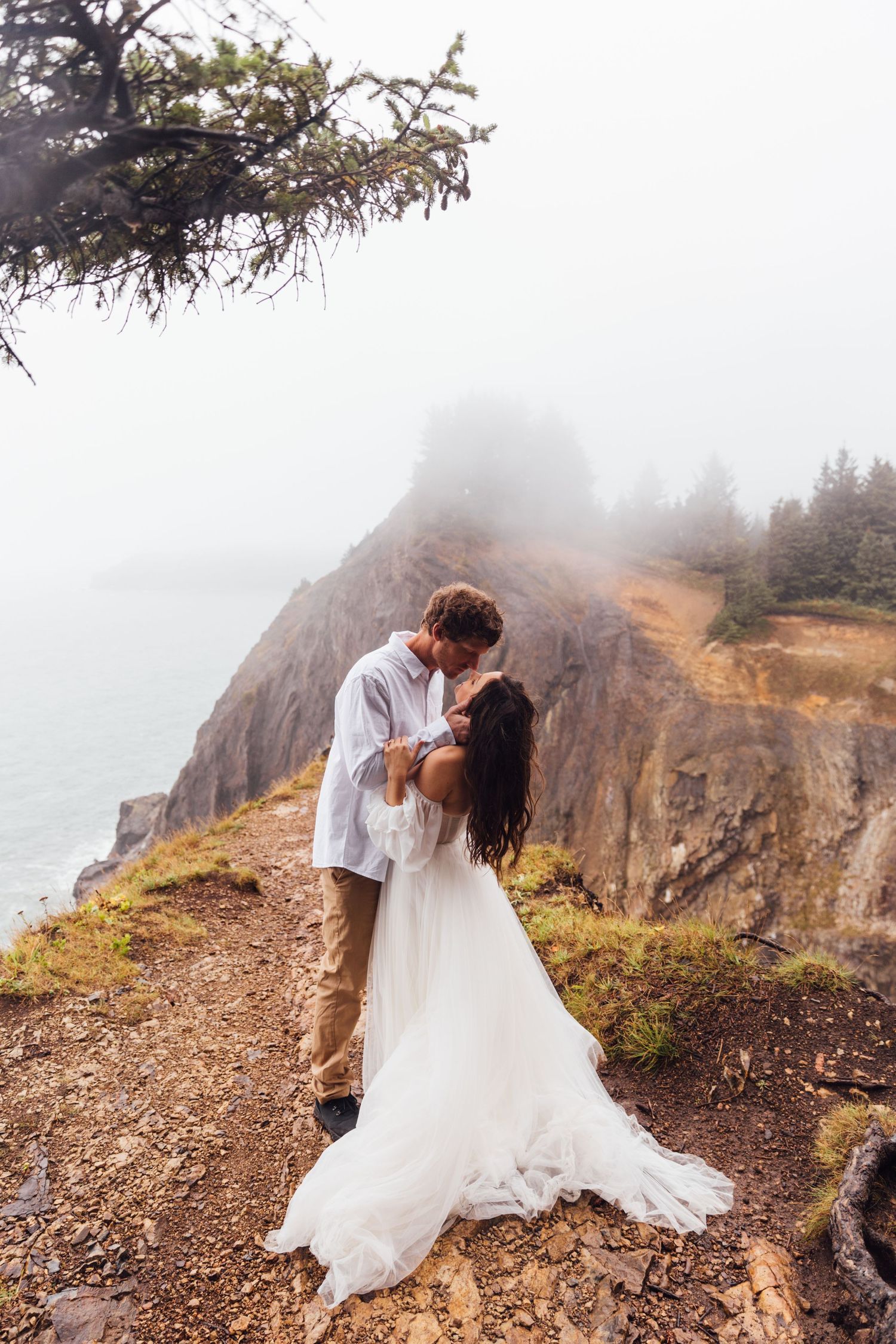 Couple in wedding attire embracing on misty mountain cliff.