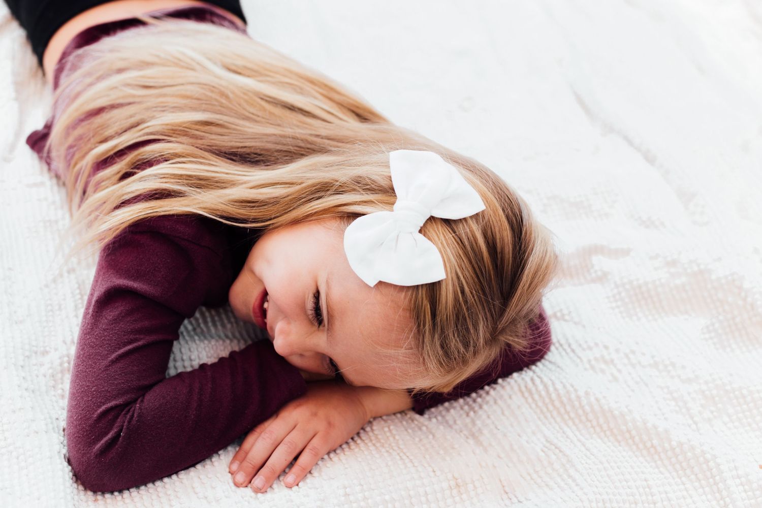 Overhead view of someone in a burgundy sweater lying on white bedding with a hair bow.