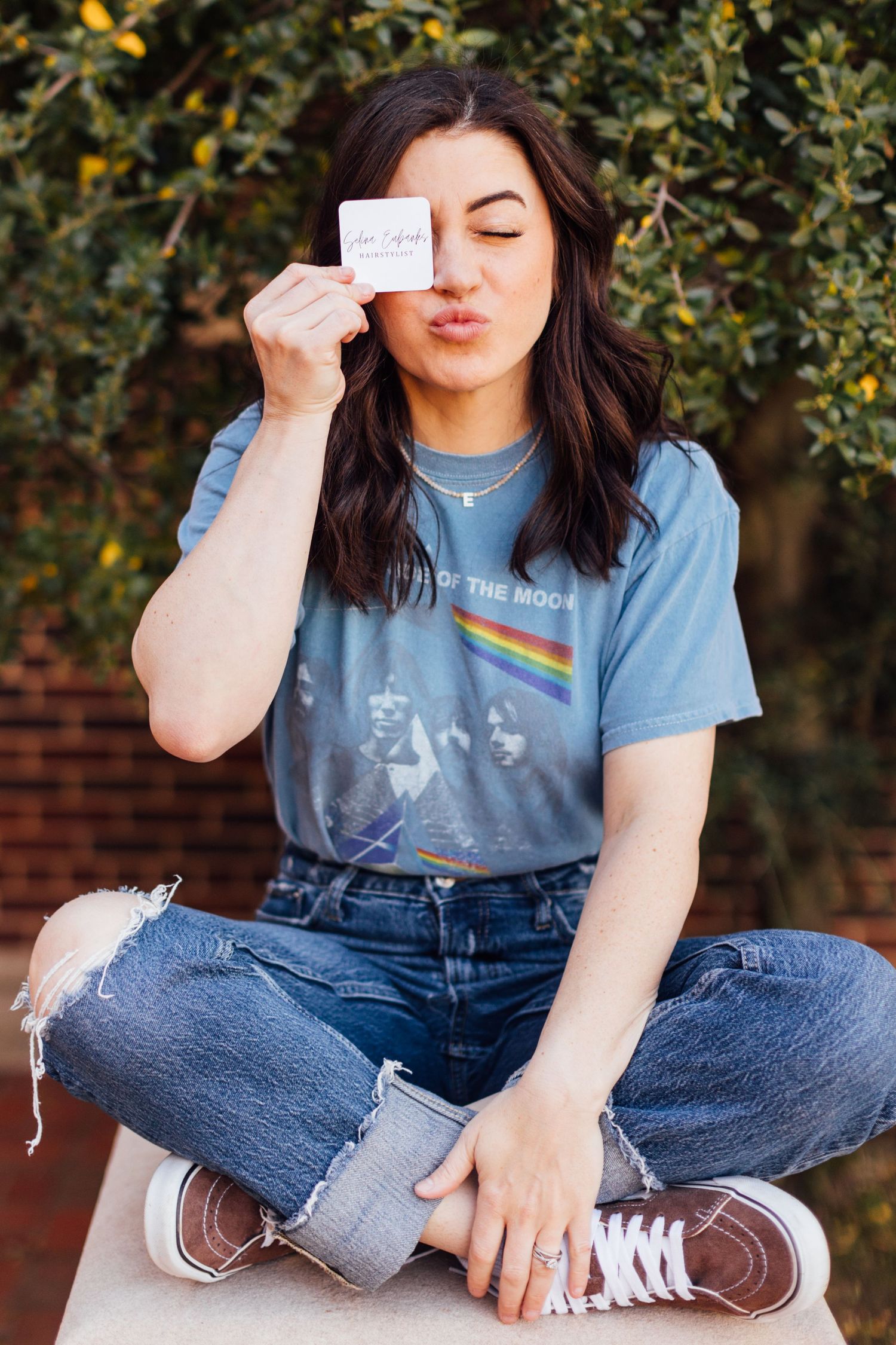 A person in overalls and a light blue t-shirt sits cross-legged outside near leafy trees and a brick wall.