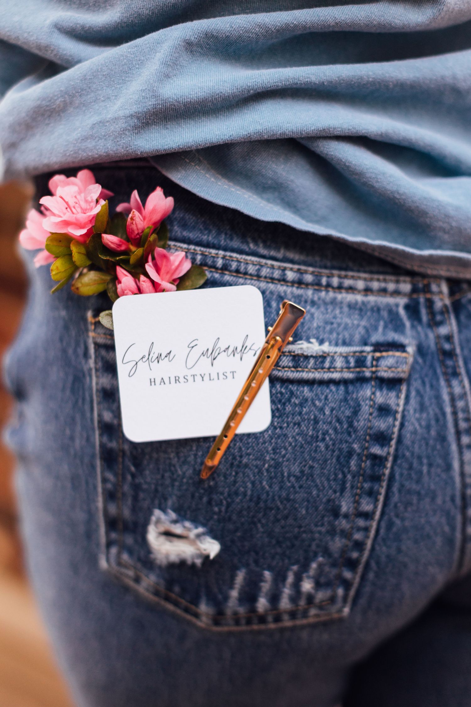 A business card tucked into the back pocket of blue jeans with delicate pink flowers for decoration.