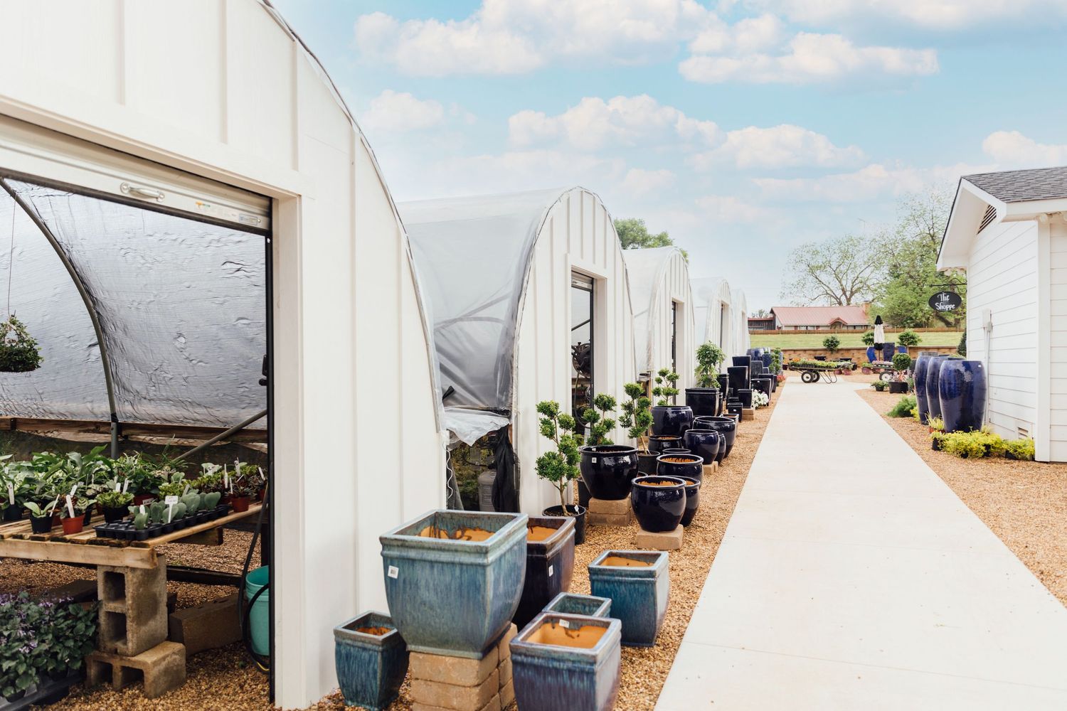 Row of white greenhouse tunnels with potted plants and containers on tables at an outdoor gardening facility in daylight.