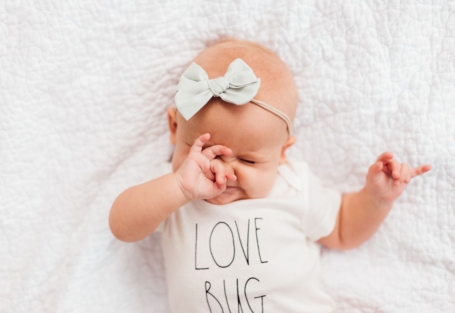 Smiling baby wearing white onesie with 'Love' text lying on white blanket with lace bow headband.