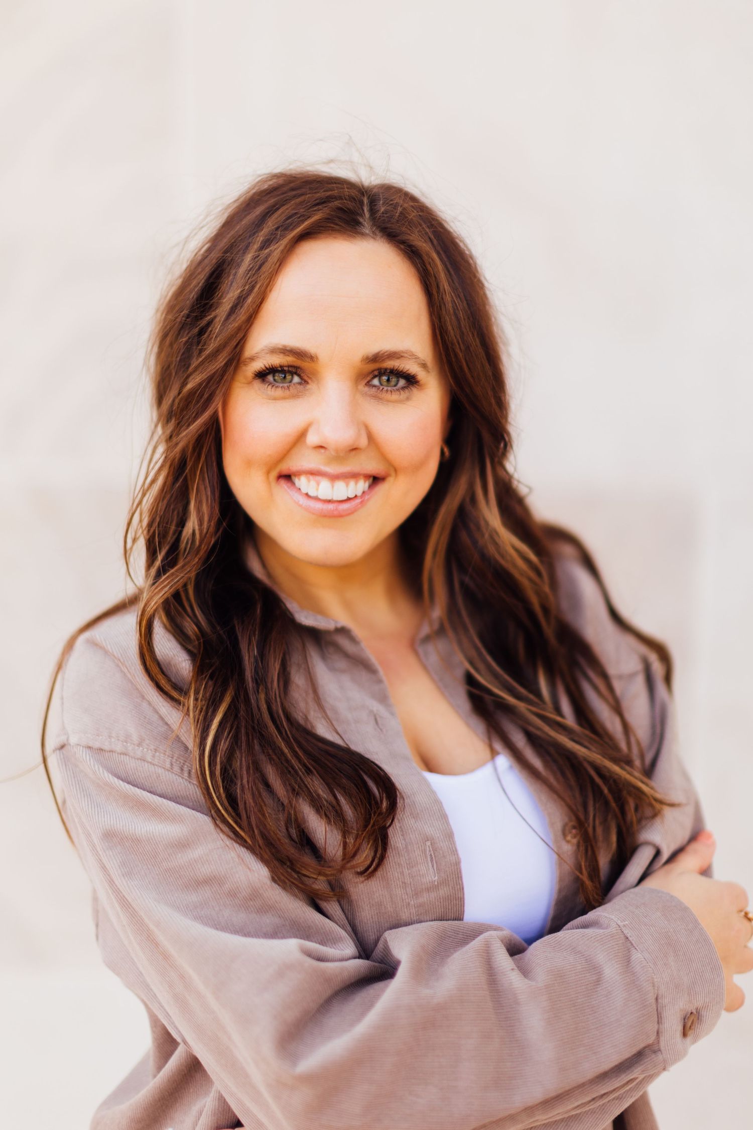 Professional portrait of someone with long brown wavy hair wearing a white top and beige jacket against a light background.