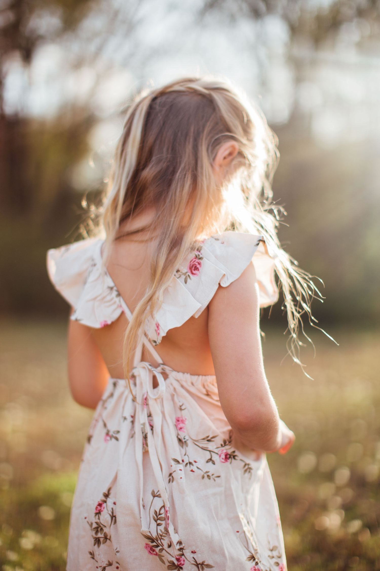 A child in a white floral dress with ruffled straps stands in a sunlit meadow during golden hour.