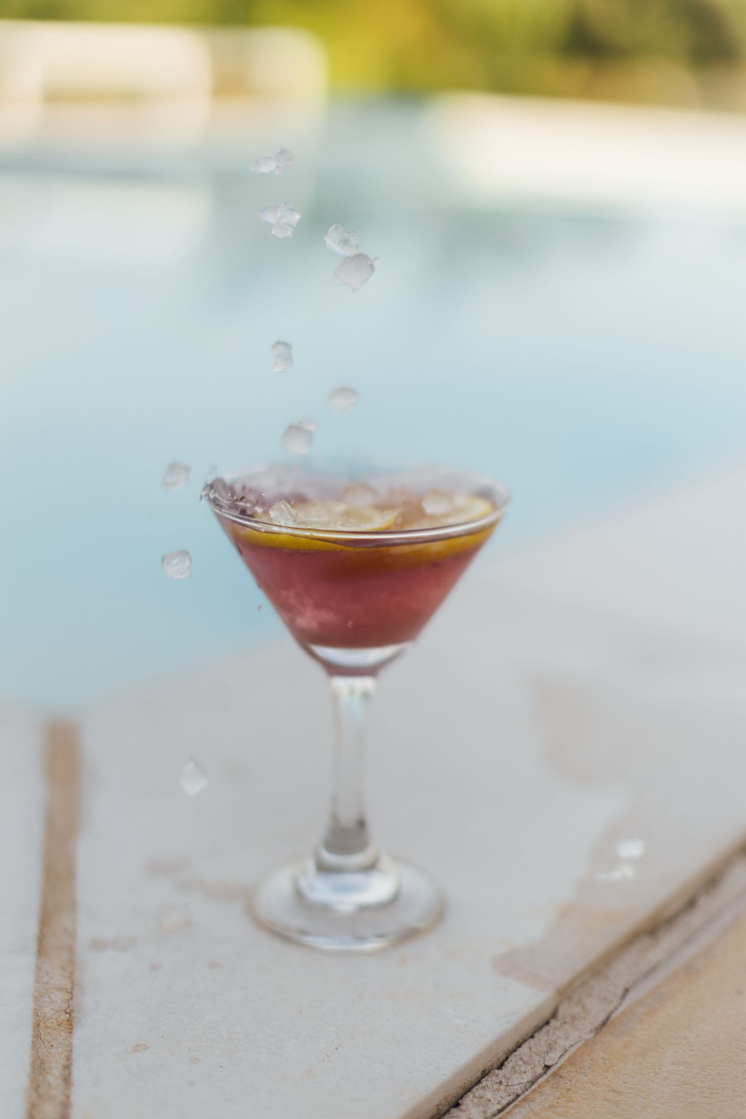 A reddish cocktail in a martini glass sits by a pool with water droplets splashing above it.