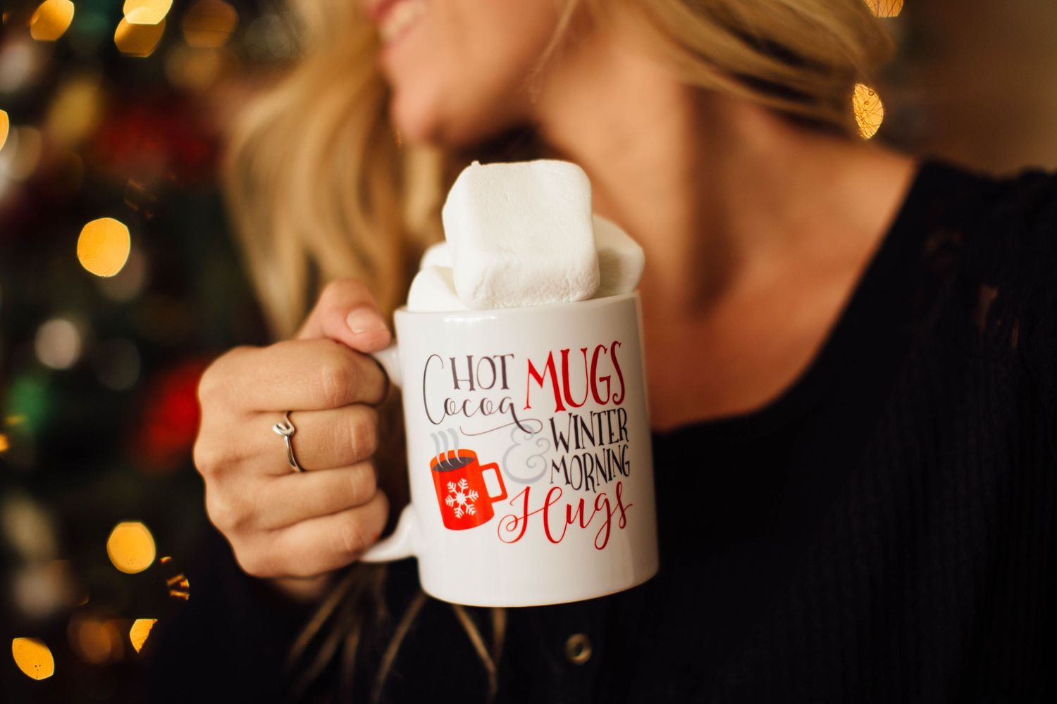 Person holding white ceramic coffee mug with Christmas text against blurred holiday lights background.
