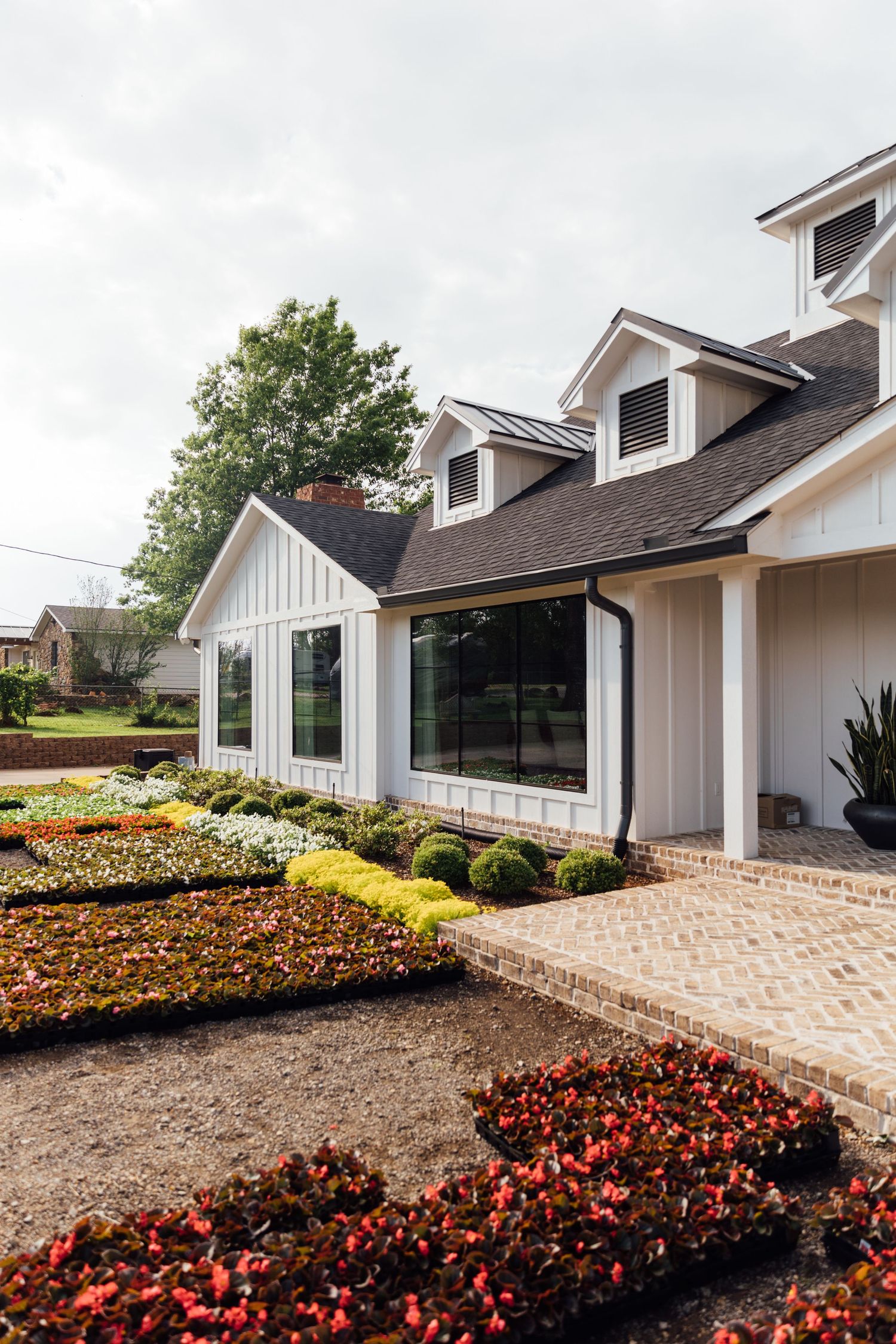 Modern white farmhouse exterior with dormer windows surrounded by vibrant red and yellow flower beds in the landscaping.