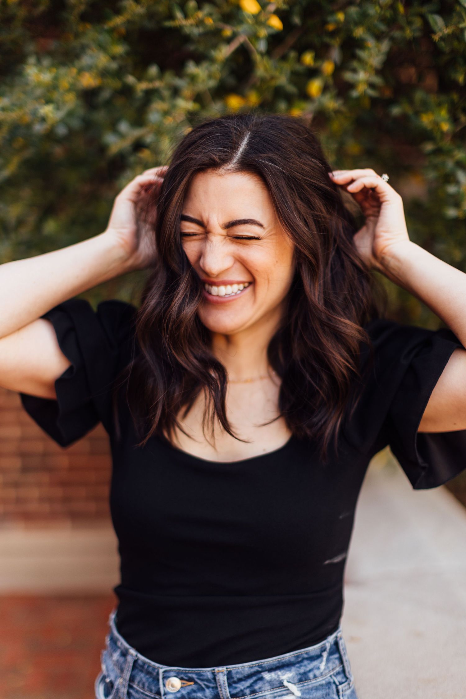 A joyful expression radiates as hands run through wavy dark hair while wearing a black top and jeans against brick backdrop.