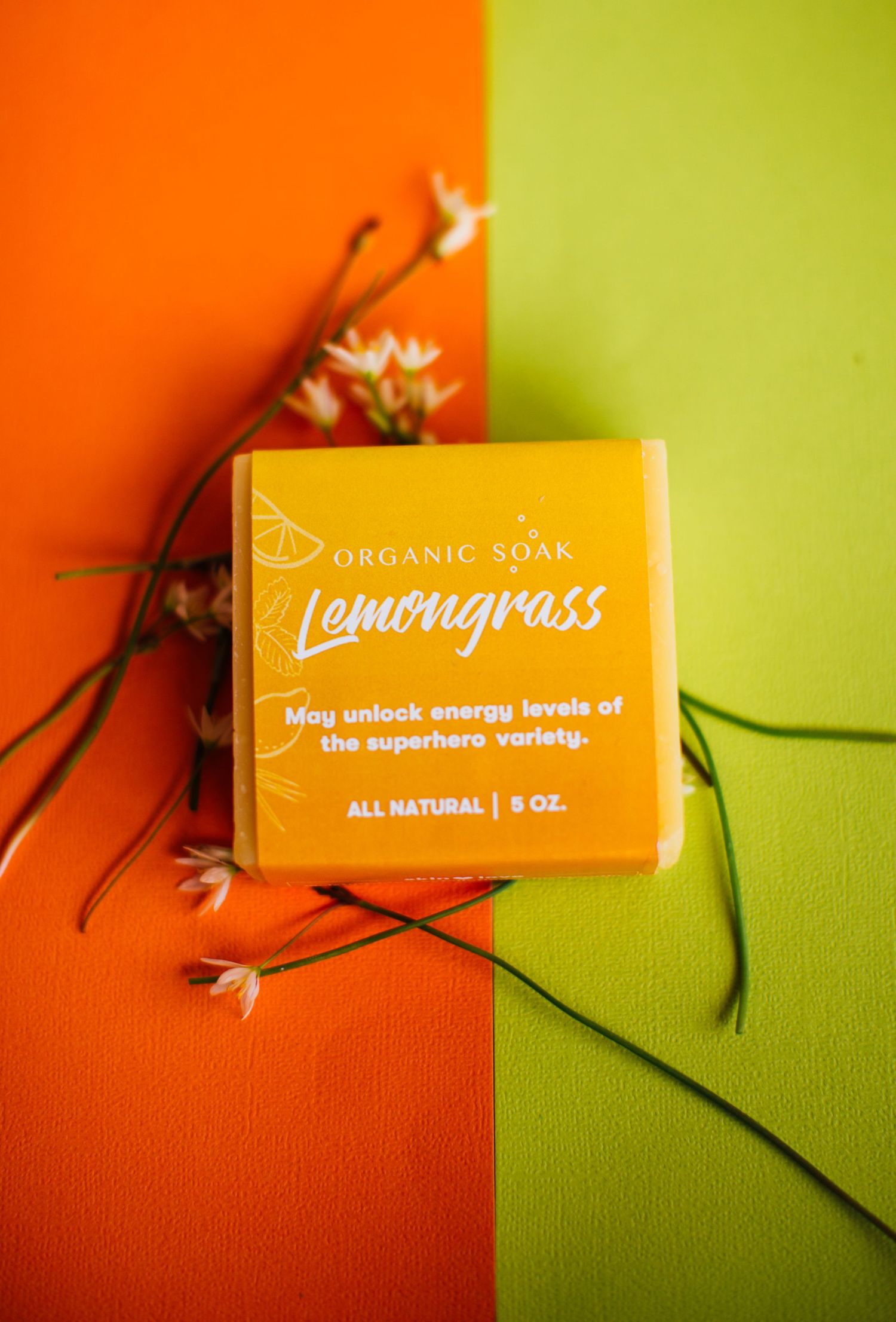 An orange lemongrass organic soap box against split orange and green background with delicate white flowers.