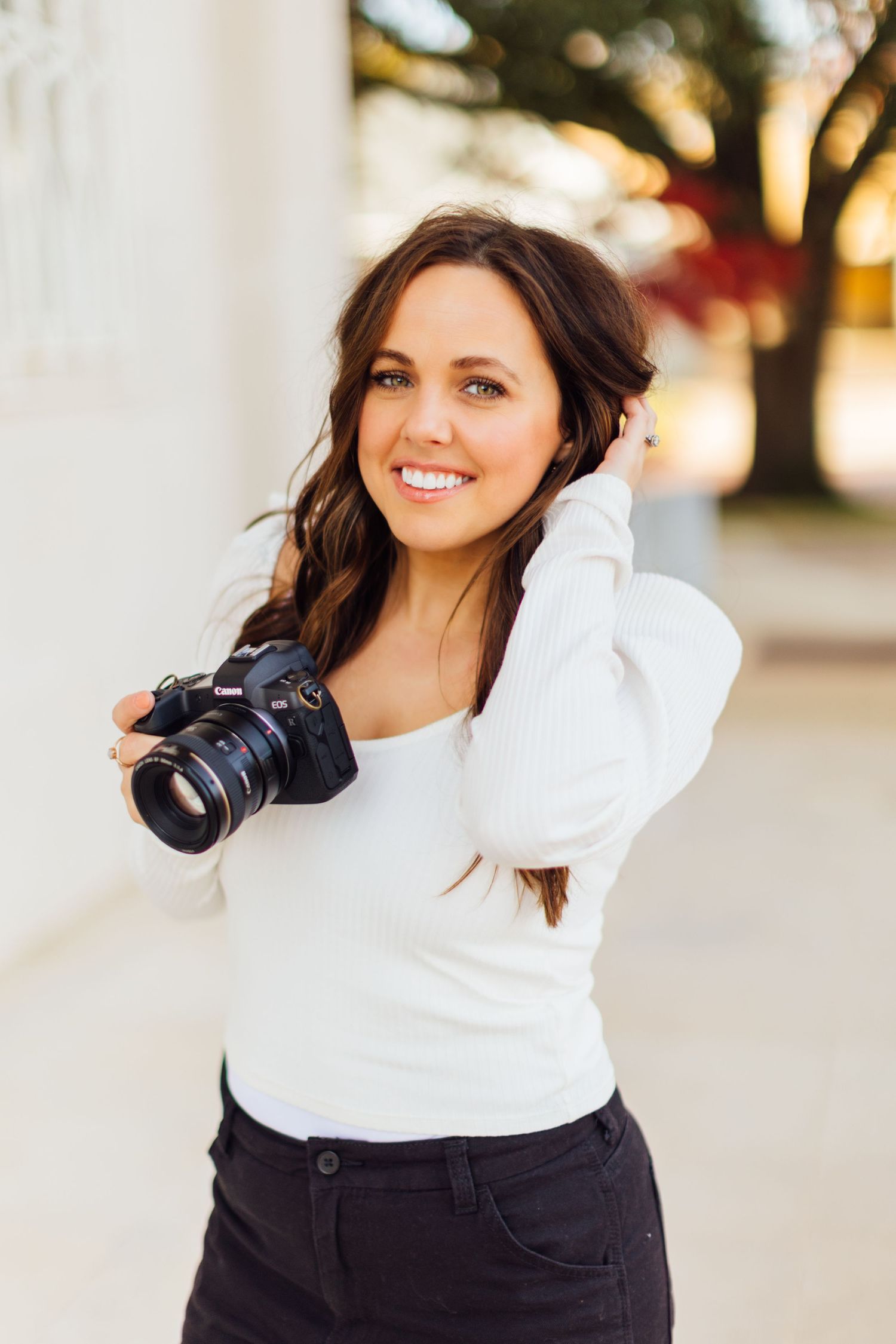 Professional photographer wearing white sweater holding DSLR camera outdoors during golden hour sunshine.