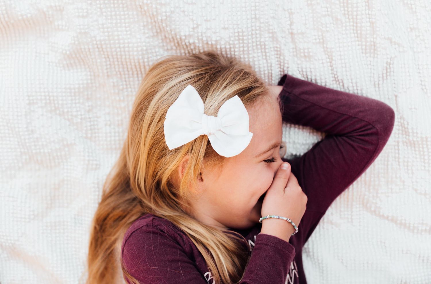 Person wearing a burgundy sweater lies on white bedding with a large white bow accessory in their hair.