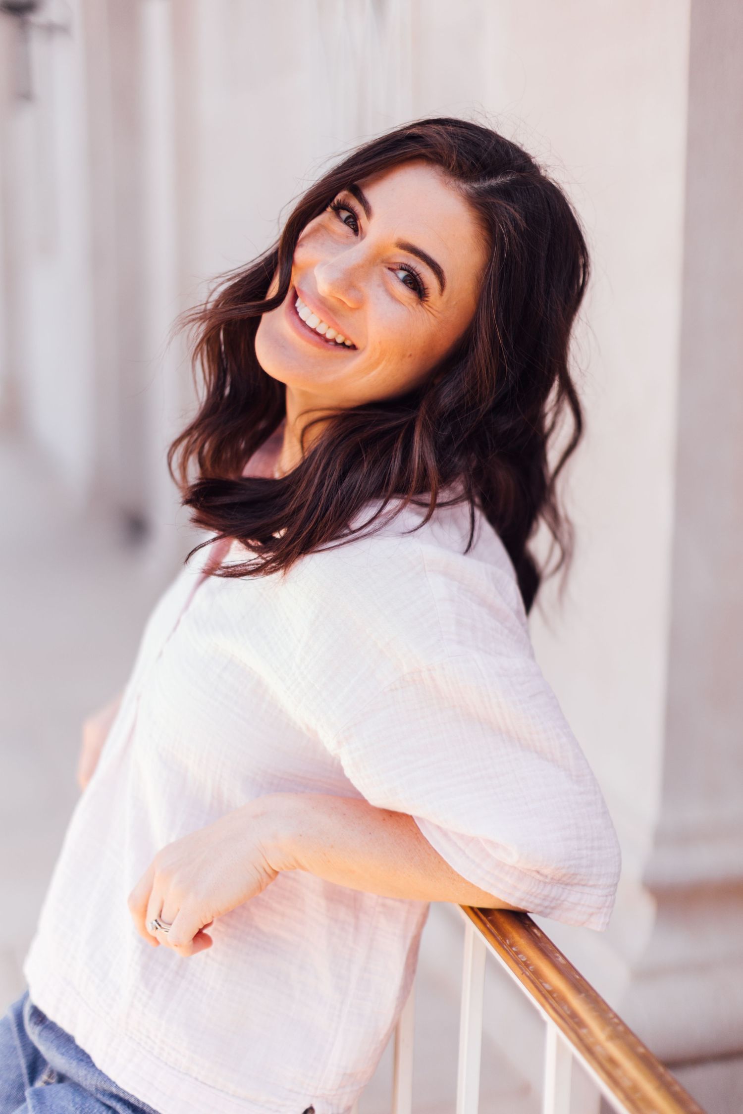 Smiling person in a white top leans against a railing while posing casually outdoors.