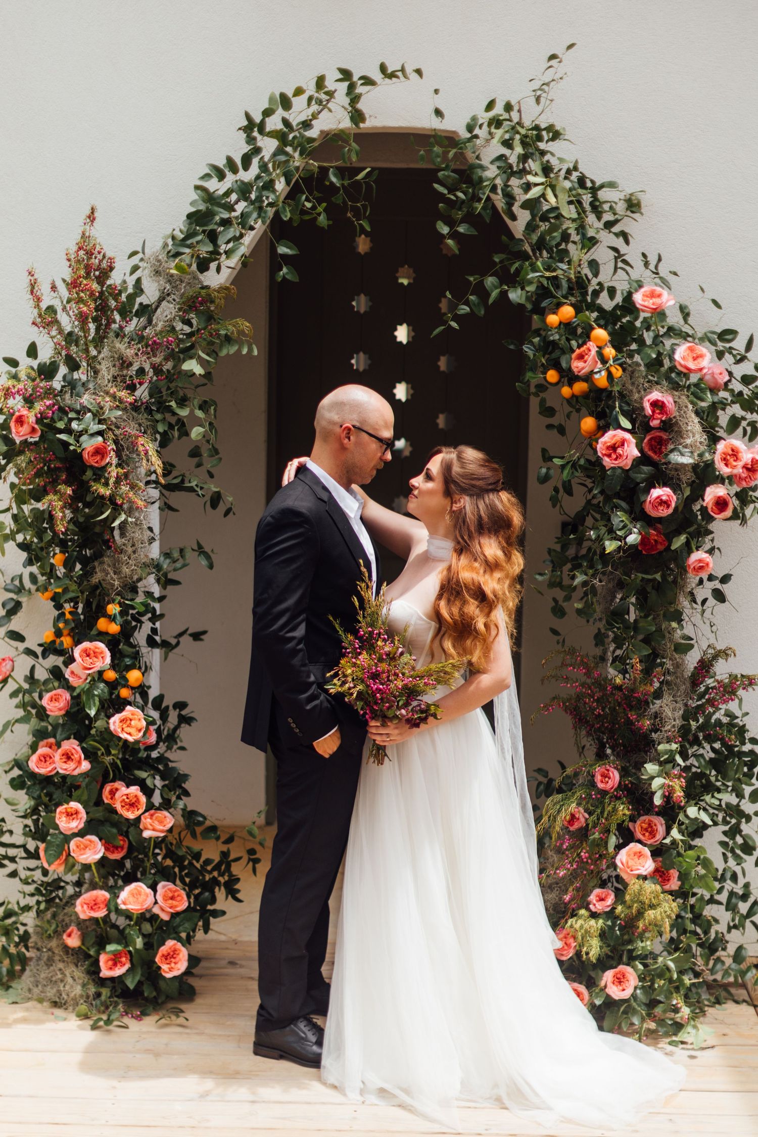 Romantic wedding portrait under floral arch with pink roses and greenery.