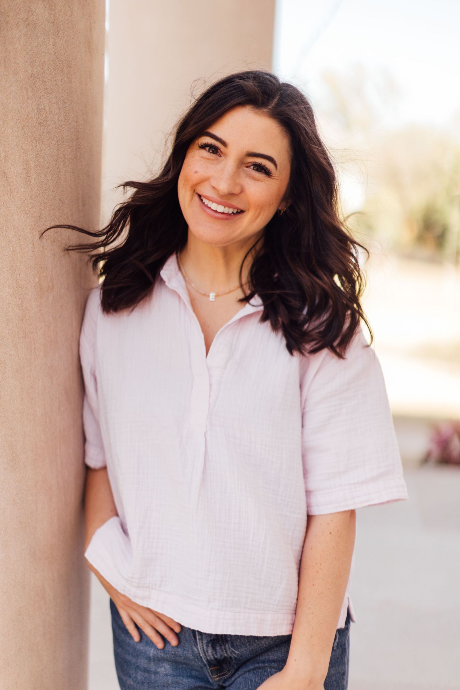 Person in light pink collared shirt and jeans leaning against beige wall outdoors while smiling warmly.