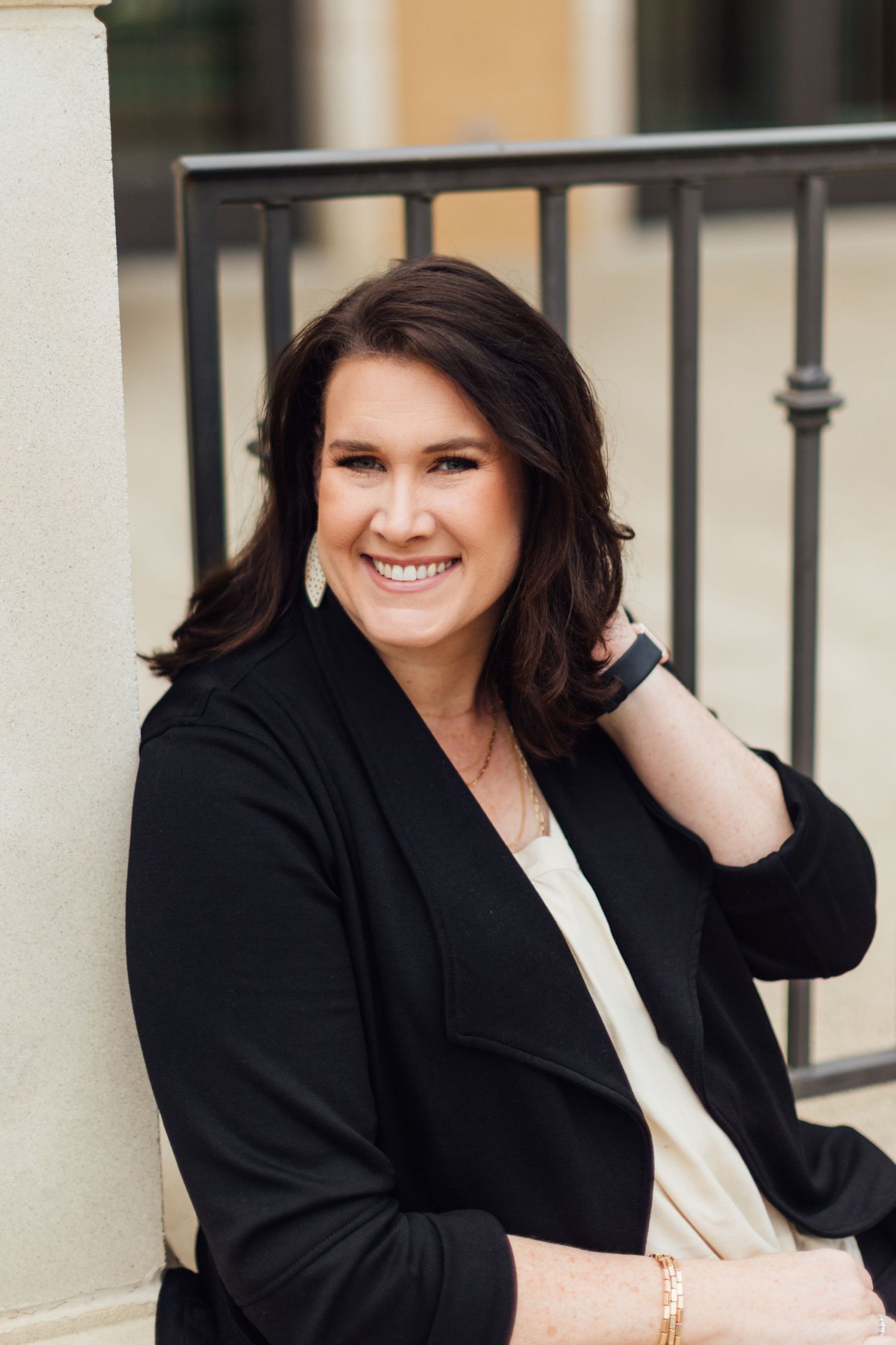 Professional portrait of a businesswoman in a black blazer smiling warmly while sitting outdoors near a railing.