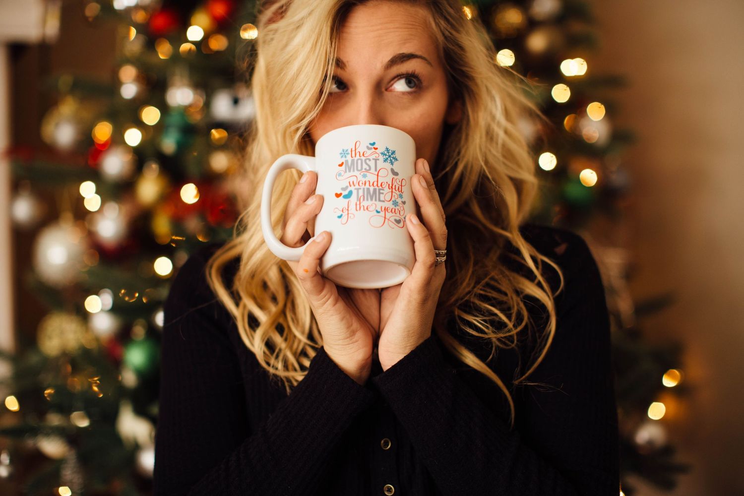 Person with wavy blonde hair holds white coffee mug while sitting near Christmas tree with bokeh lights in background.