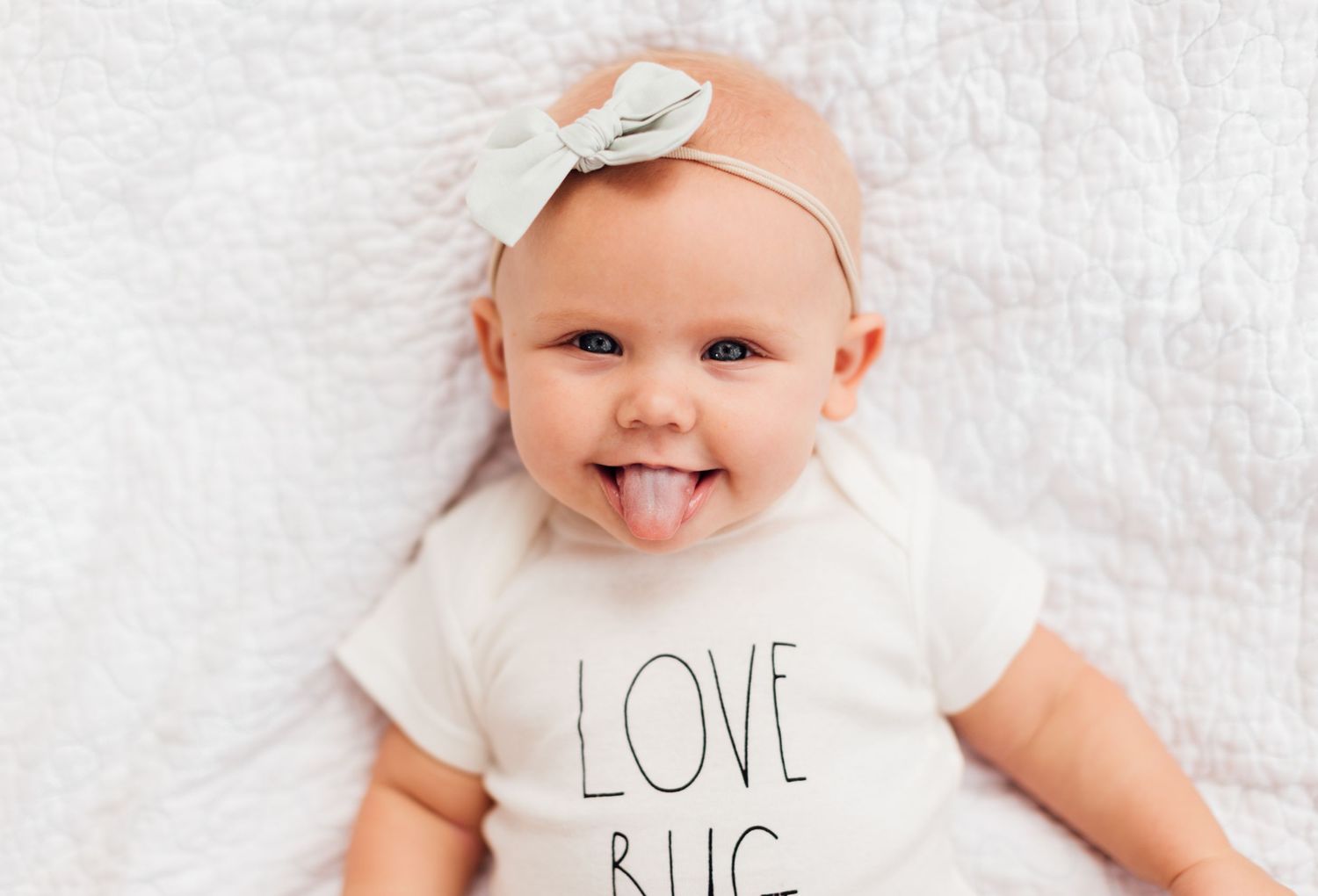 Baby wearing white onesie with text lying on white blanket smiles at camera.