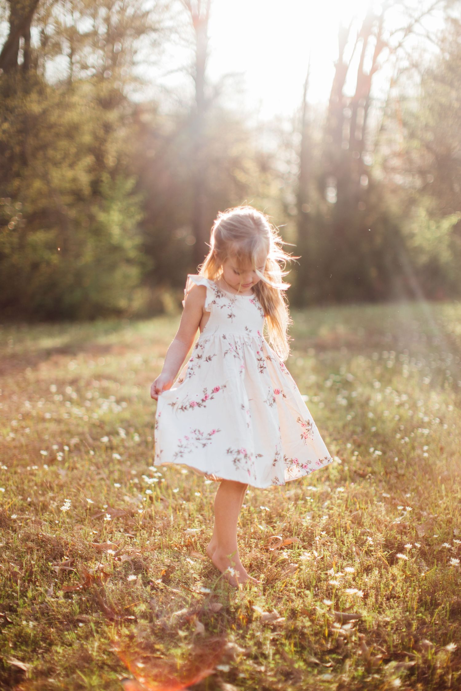 Child in white floral dress twirls and dances in a sunlit meadow during golden hour.