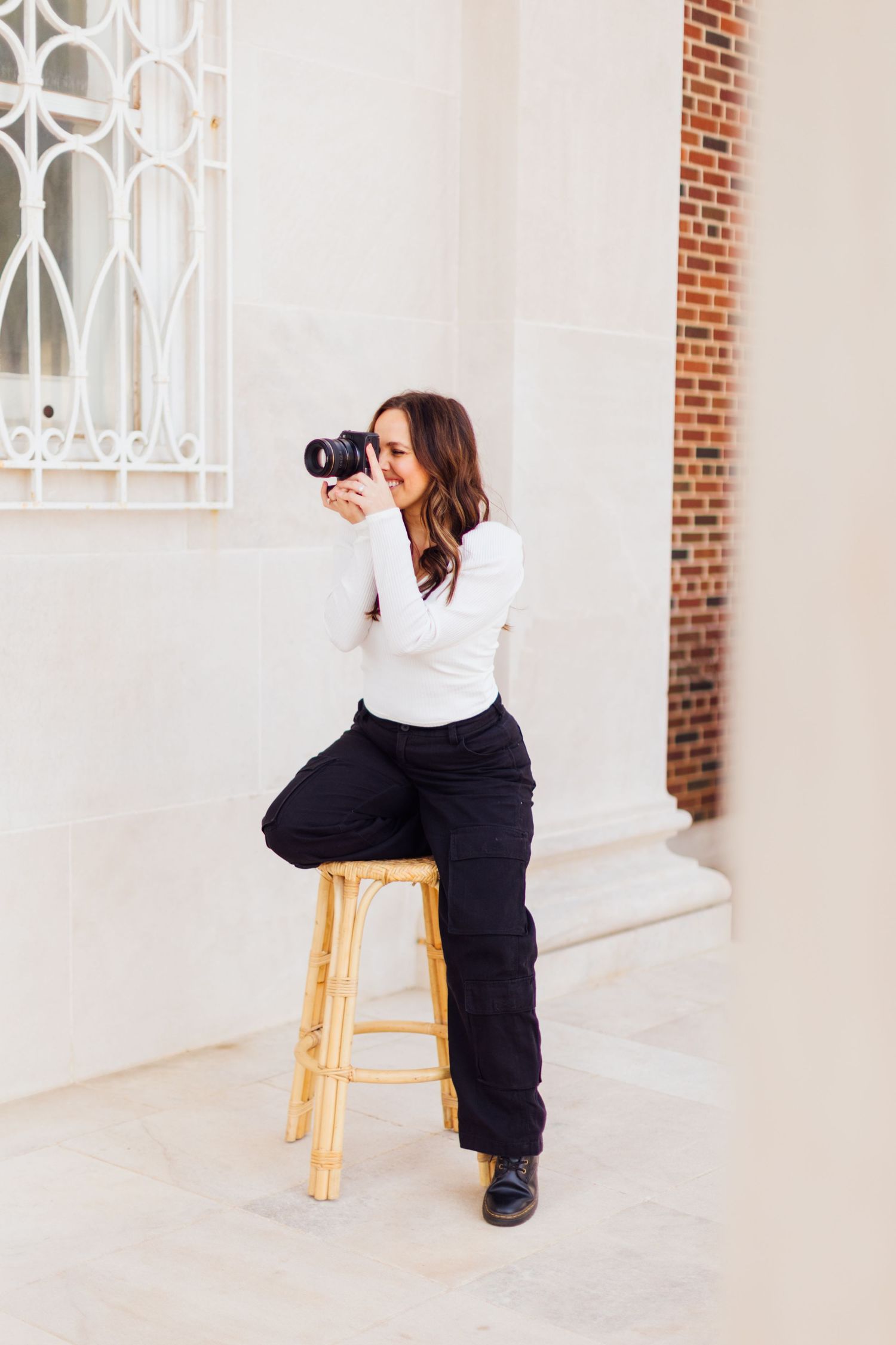 A photographer wearing a white shirt and black pants sits on a wooden stool while taking pictures with a professional camera.