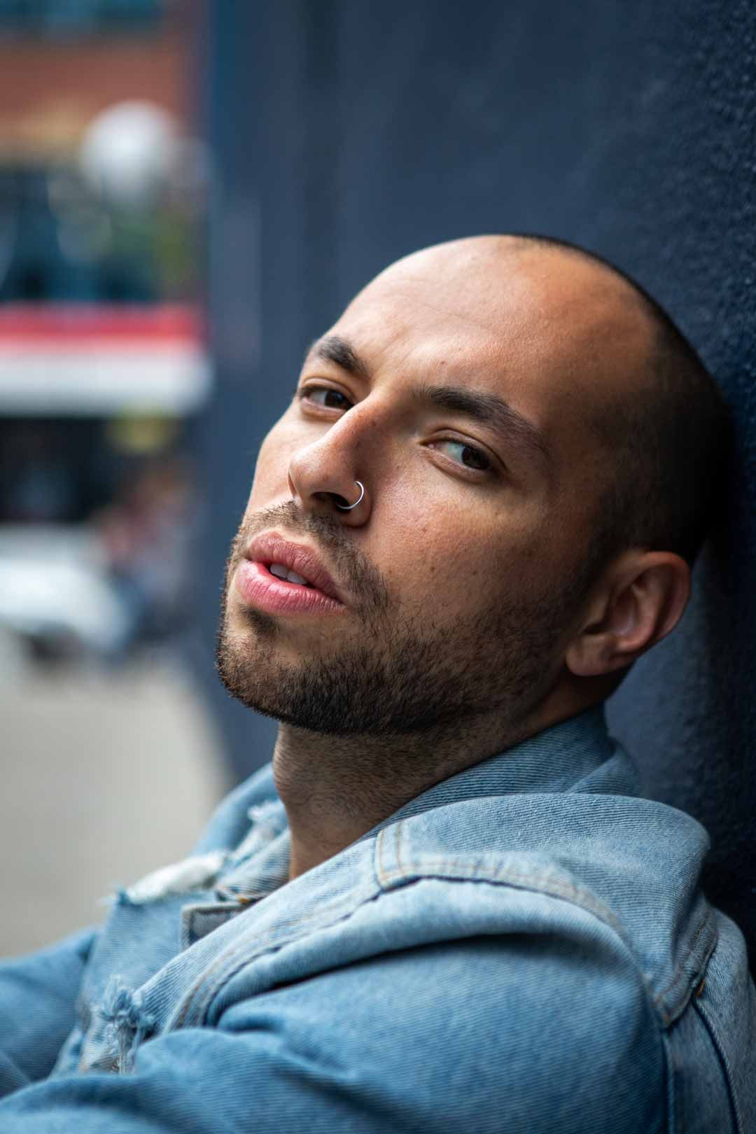 Professional headshot of bald man with beard and nose ring in denim jacket on urban street
