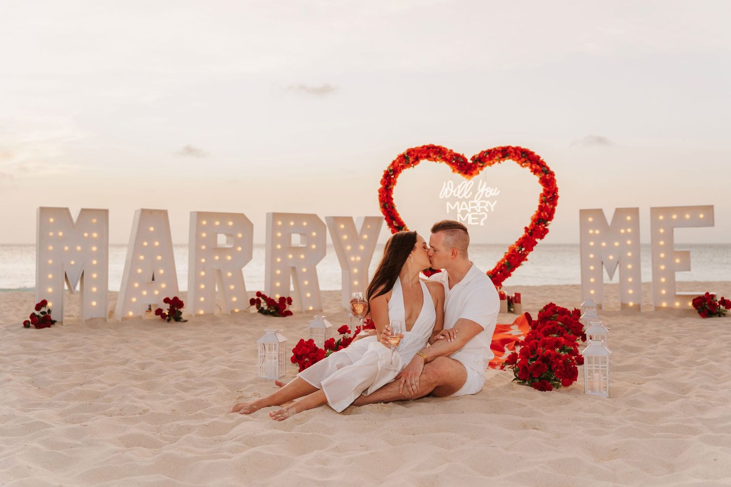 A romantic beach proposal setup with illuminated 'MARRY ME' letters, a heart arch, and red roses on the sand at dusk.