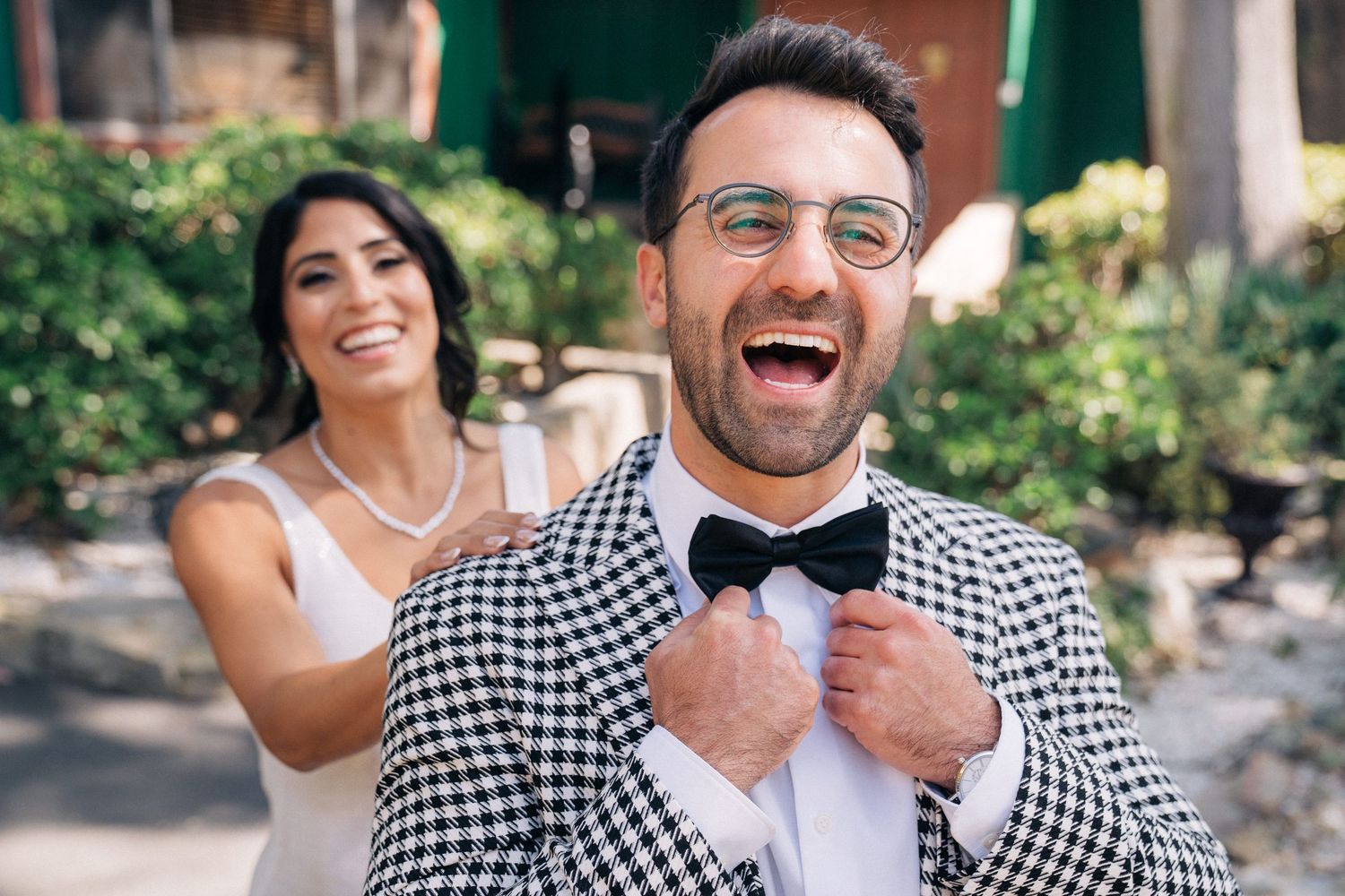 A couple in gingham attire shares candid laughter and joyful moments during an outdoor photoshoot in a garden setting.
