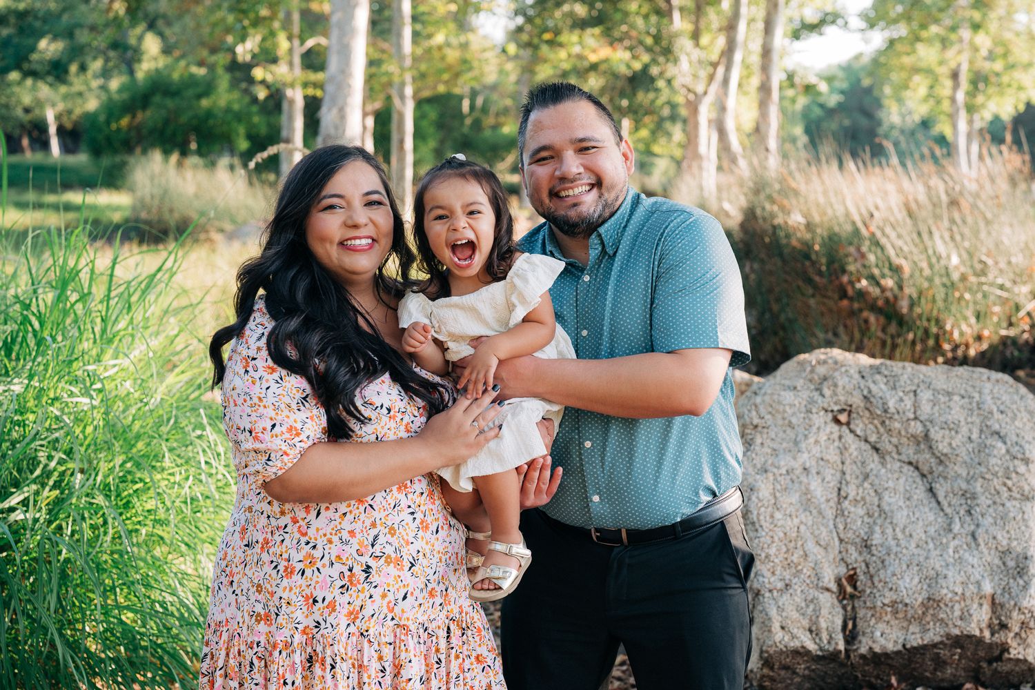 Three people and a small white dog pose together in an outdoor garden setting surrounded by lush greenery.