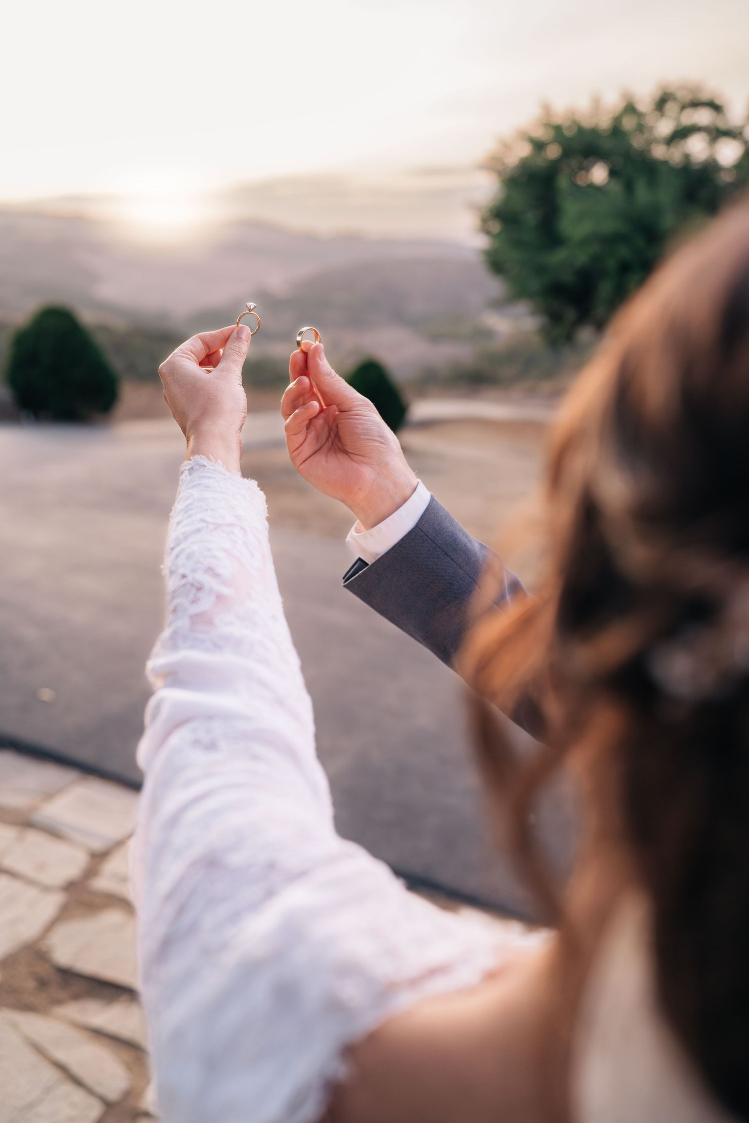 Two hands, one in a white sleeve and one in a suit sleeve, reach towards a scenic sunset view over a stone terrace.