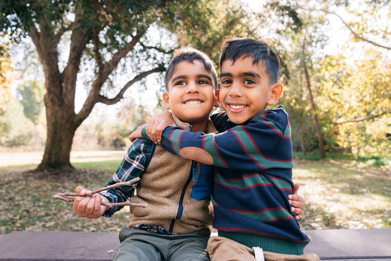 Two children in warm clothing laugh and play together outdoors during autumn among fallen leaves.