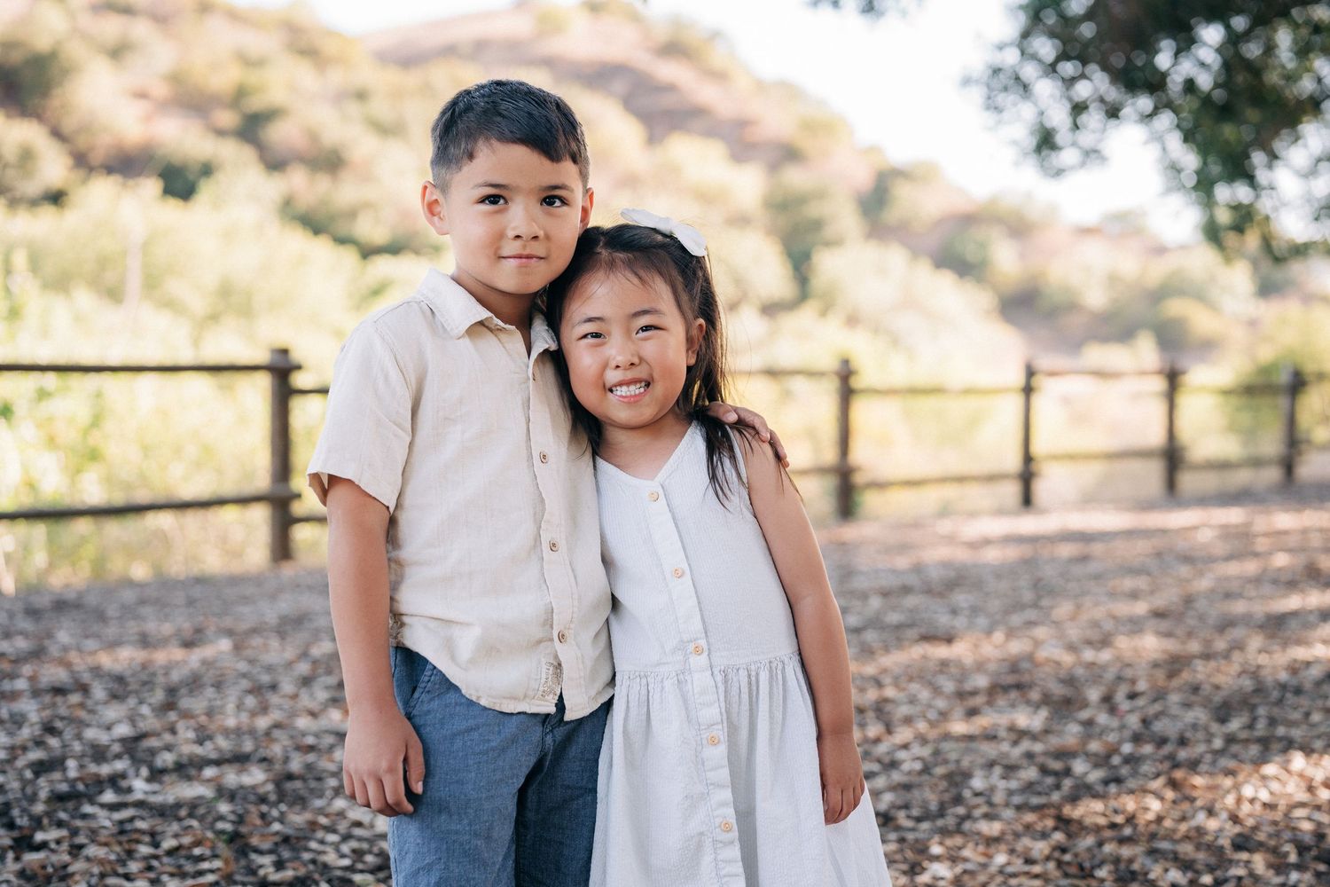 Two young children stand together smiling outdoors near a wooden fence in a natural setting.