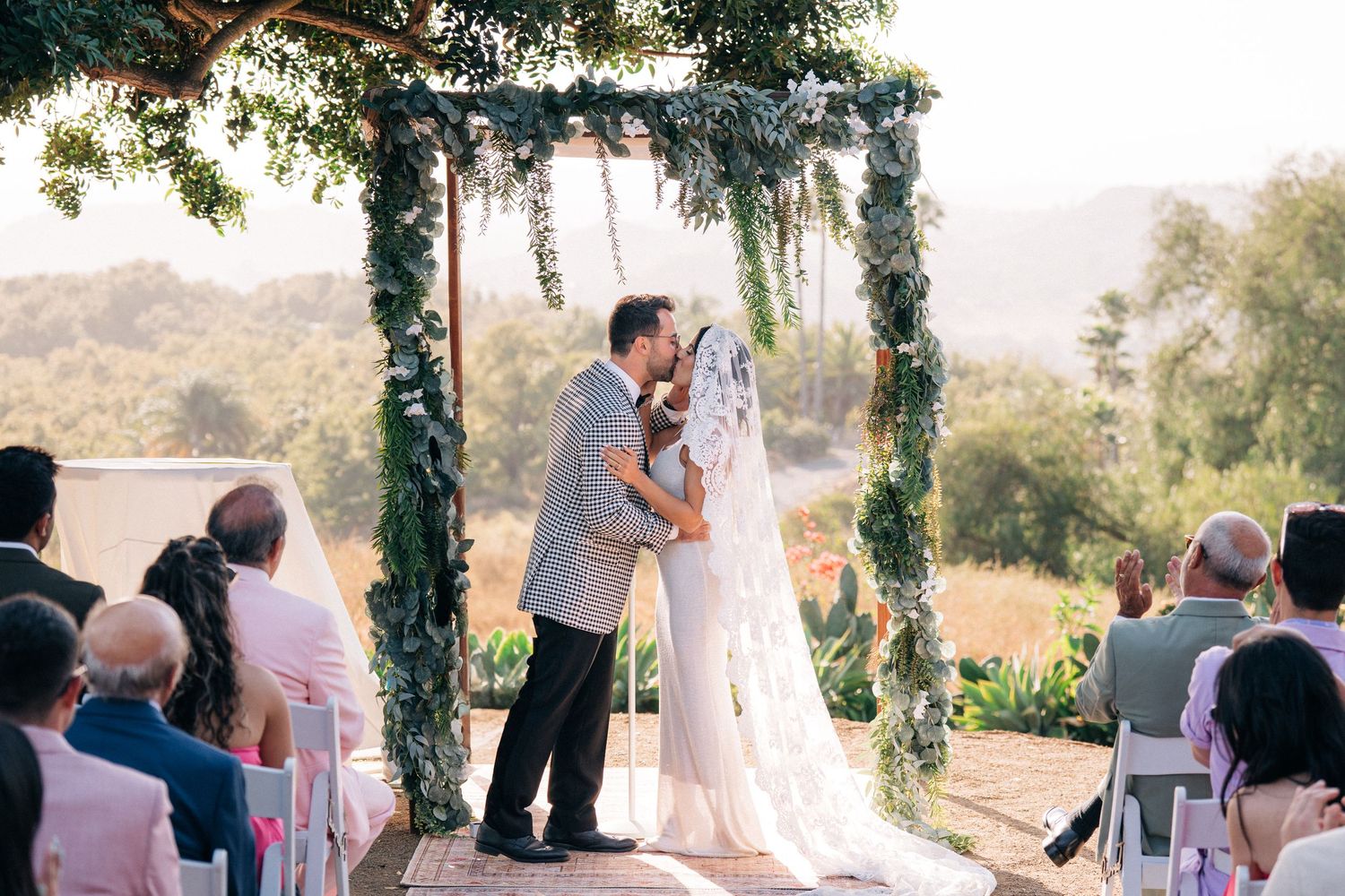 A romantic outdoor wedding ceremony under a floral arch at sunset with guests seated on chairs in a natural setting.