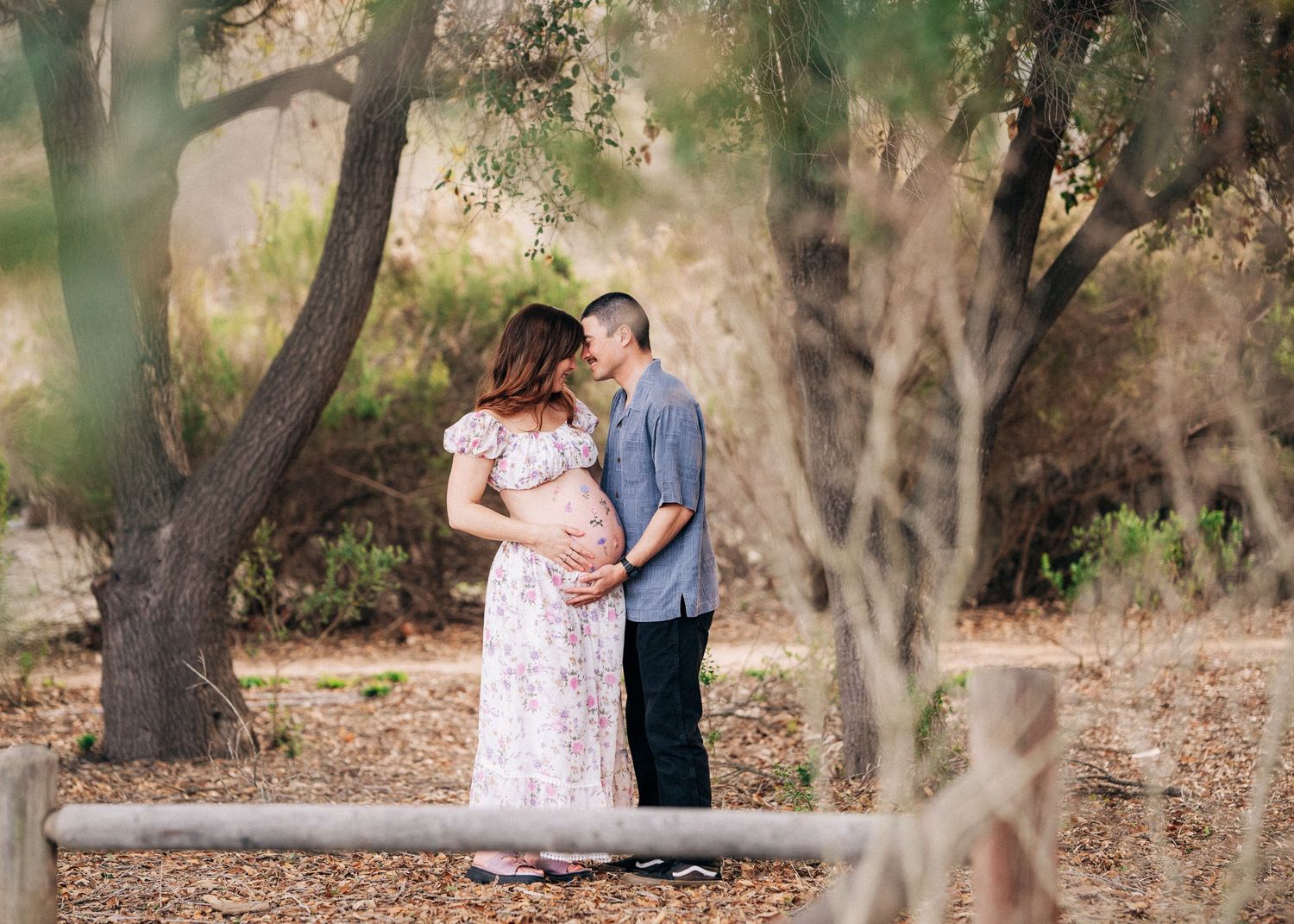 Couple embracing in a romantic outdoor setting with trees and dappled sunlight in the background.
