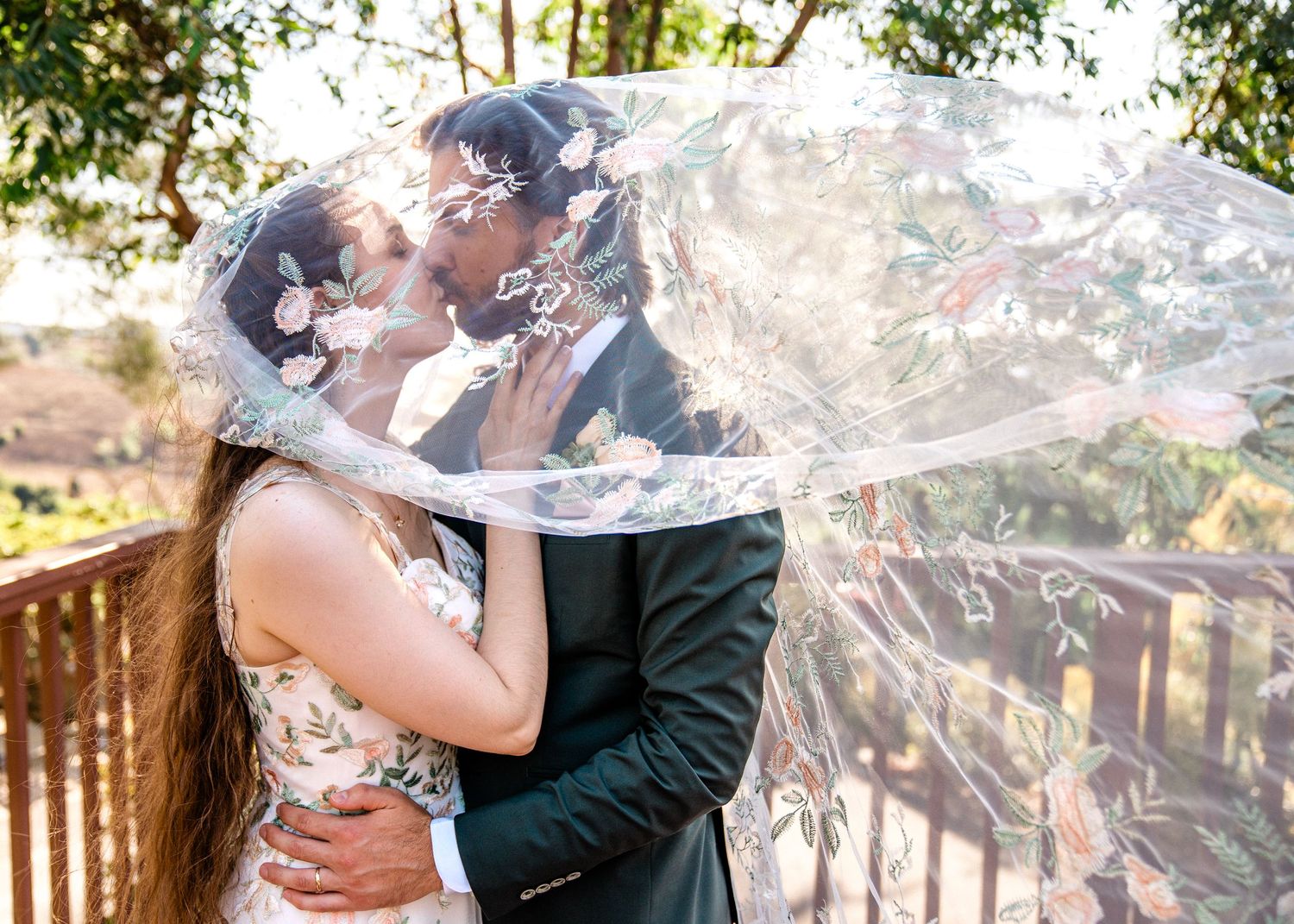 The couple shares an intimate embrace and kiss on a wooden deck surrounded by sunlit trees.
