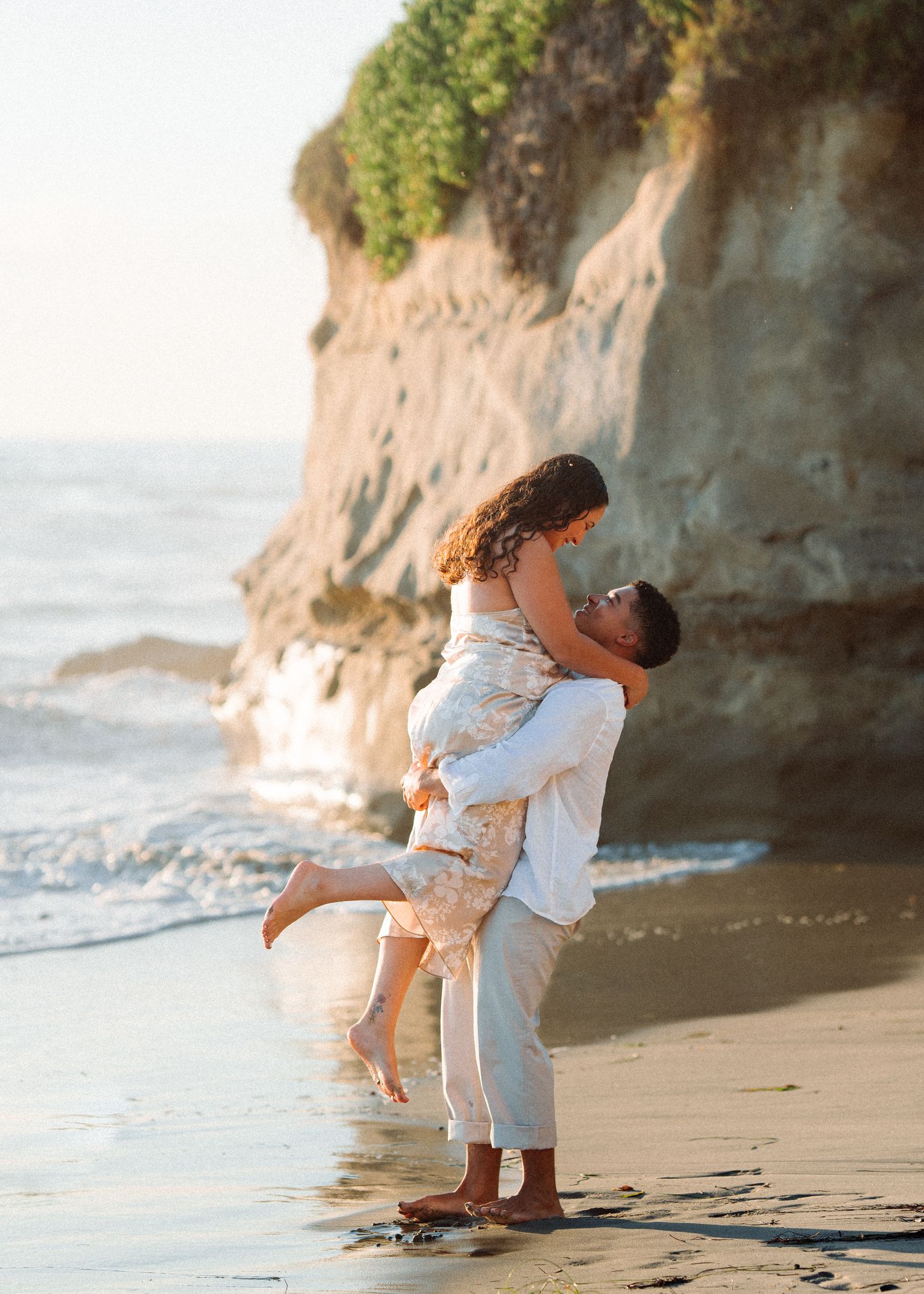 Romantic couple embraces on scenic beach at sunset with rocky cliffs in the background.