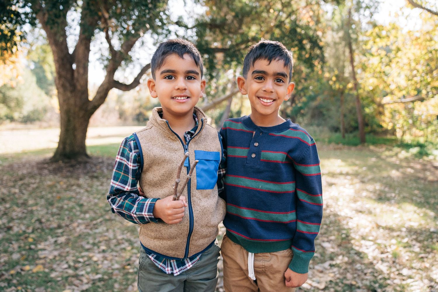 Two young children in casual clothing playing and laughing together in a sunlit wooded outdoor setting.